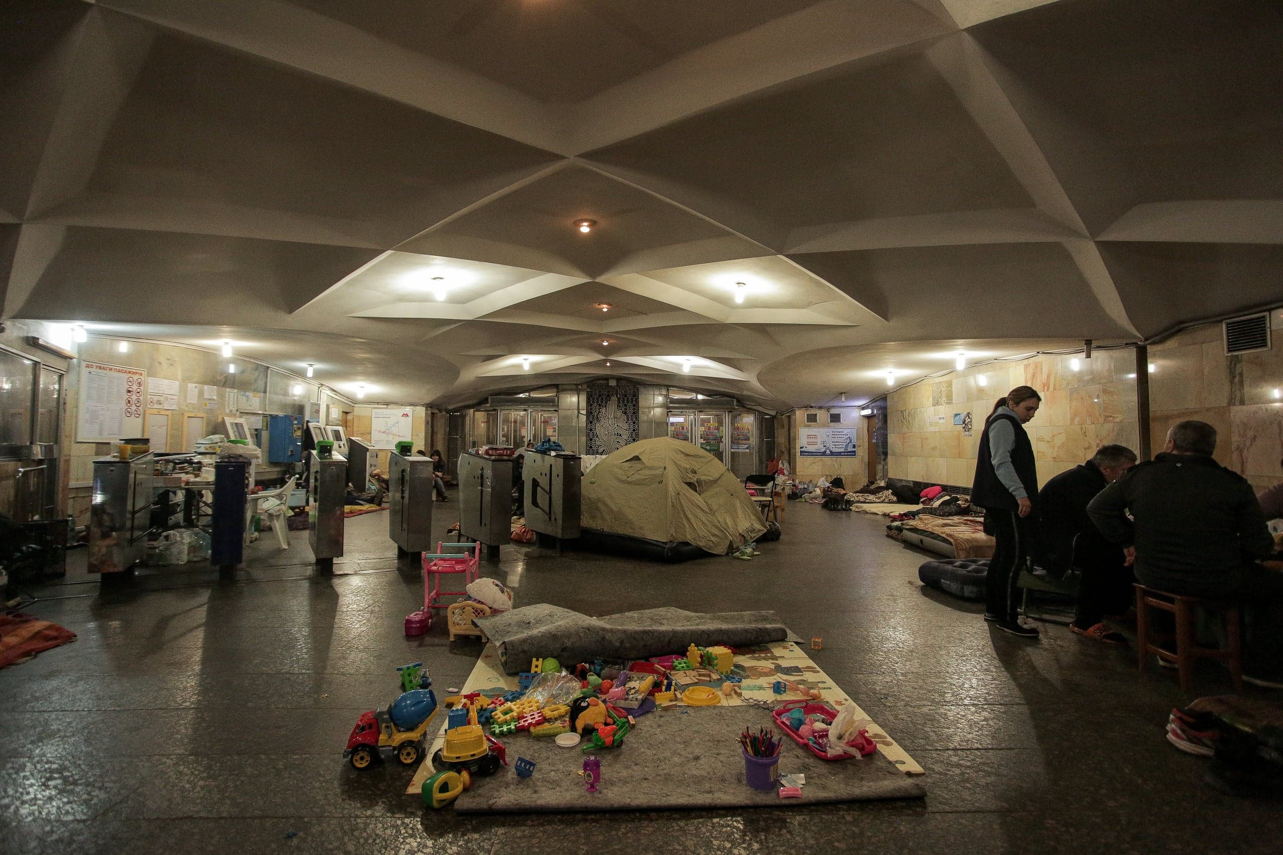 Зал станции метро, используемый как убежище, с матрасами, палаткой, турникетами и детскими игрушками на полу.