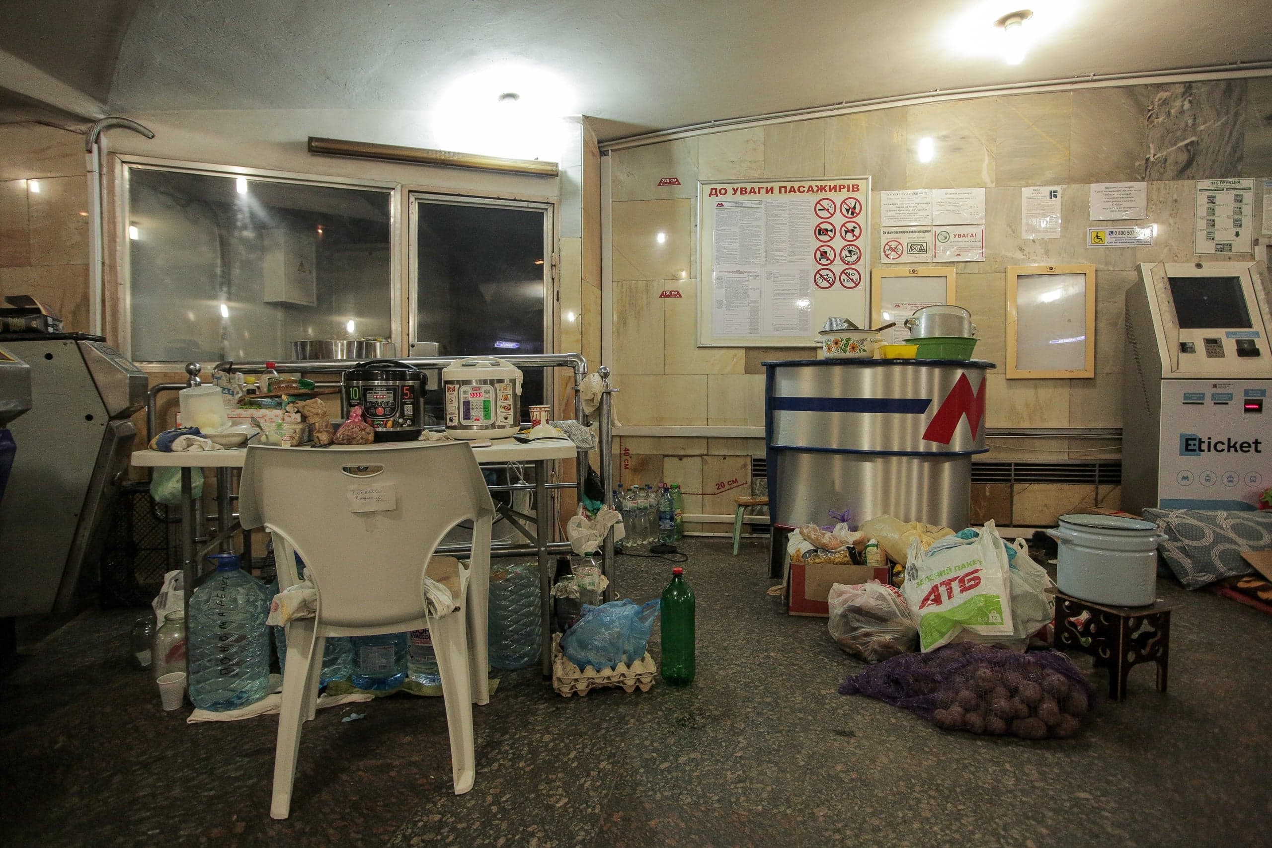 Интерьер зала ожидания, переоборудованный под временное жилье. Стол с кухонными приборами и едой, пластиковый стул, мешки с продуктами и водой на полу. На стене плакат "ДО УВАГИ ПАСАЖИРІВ" и автомат "Eticket".