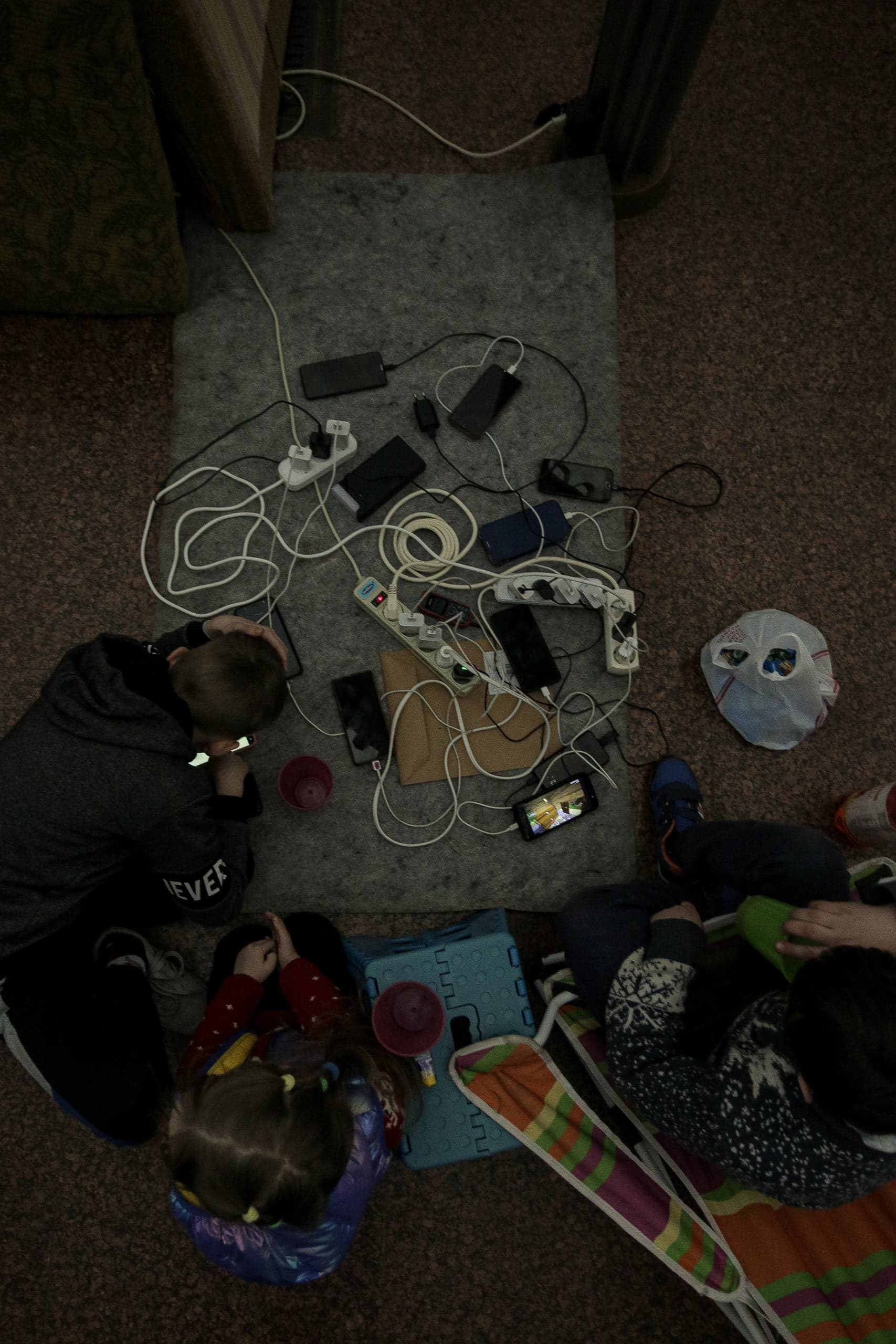 Группа людей сидит на полу в полумраке вокруг хаотично расположенных удлинителей и множества заряжающихся смартфонов.