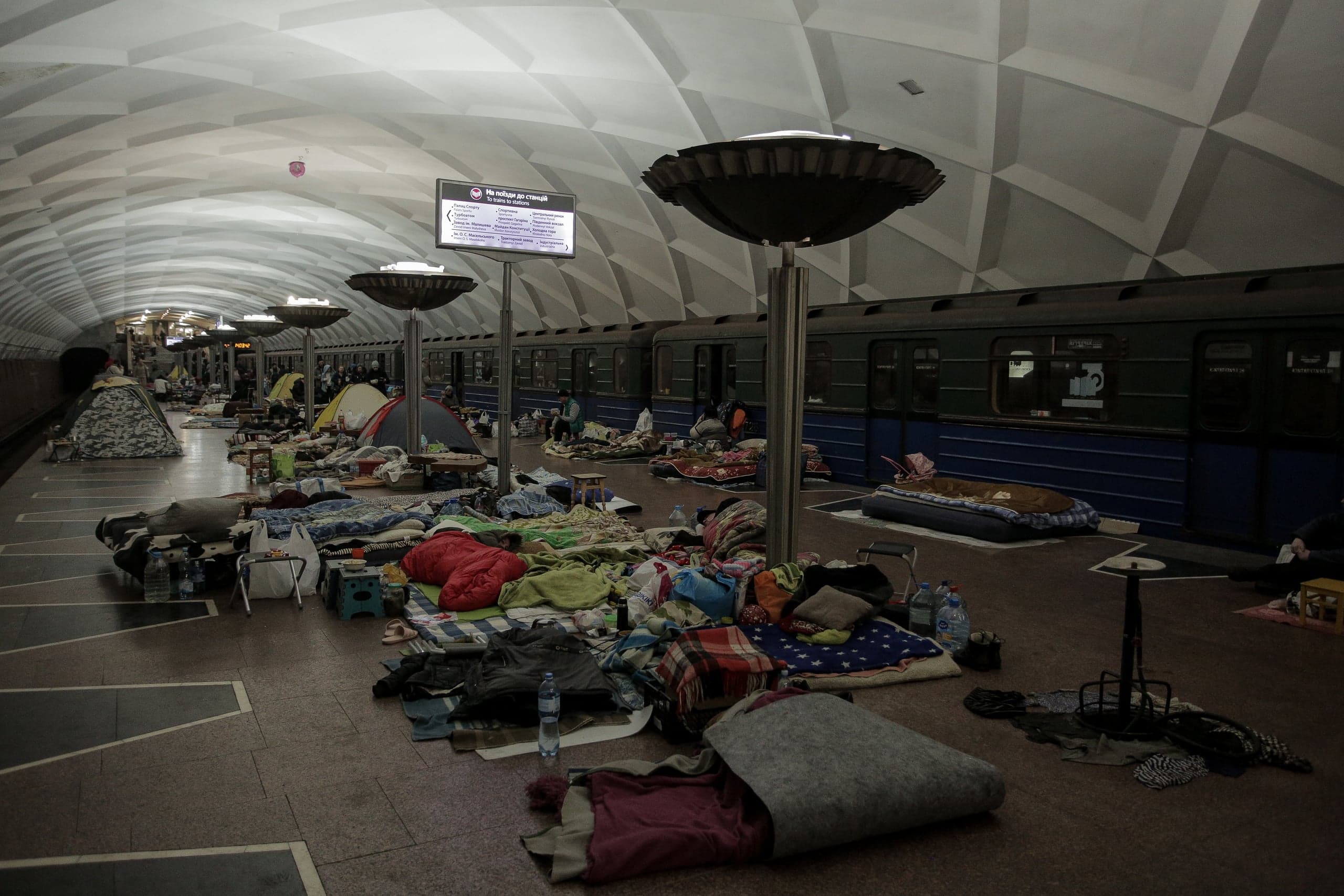 Платформа метро, используемая в качестве убежища. Пол покрыт матрасами, спальными мешками, одеялами и личными вещами. Вдоль перрона стоит поезд.