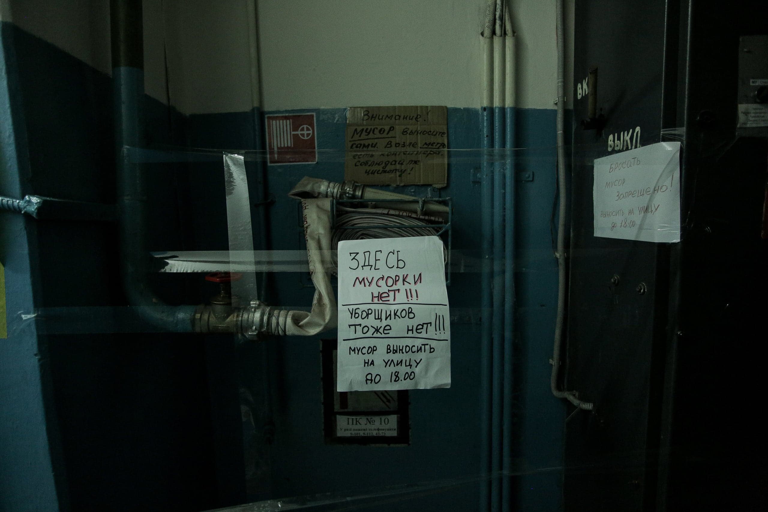 Темное служебное помещение с трубами, пожарным рукавом и тремя рукописными объявлениями. Надписи: "ЗДЕСЬ МУСОРКИ НЕТ !!! УБОРЩИКОВ ТОЖЕ НЕТ !!! МУСОР ВЫНОСИТЬ НА УЛИЦУ ДО 18.00", "ВНИМАНИЕ! МУСОР ВЫНОСИТЕ сами. Возле лифта есть контейнер. Соблюдайте чистоту!" и "БРОСАТЬ МУСОР ЗАПРЕЩЕНО! ВЫНОСИТЬ НА УЛИЦУ ДО 18.00".