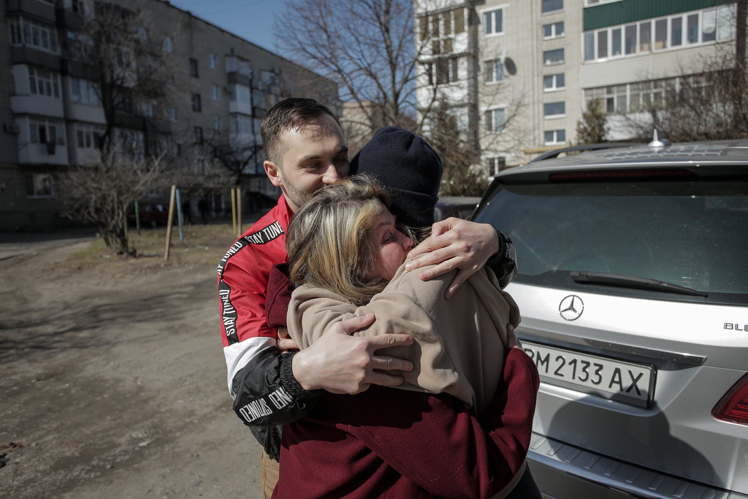 Эмоциональные объятия мужчины и двух других людей на улице на фоне жилых домов и автомобиля.