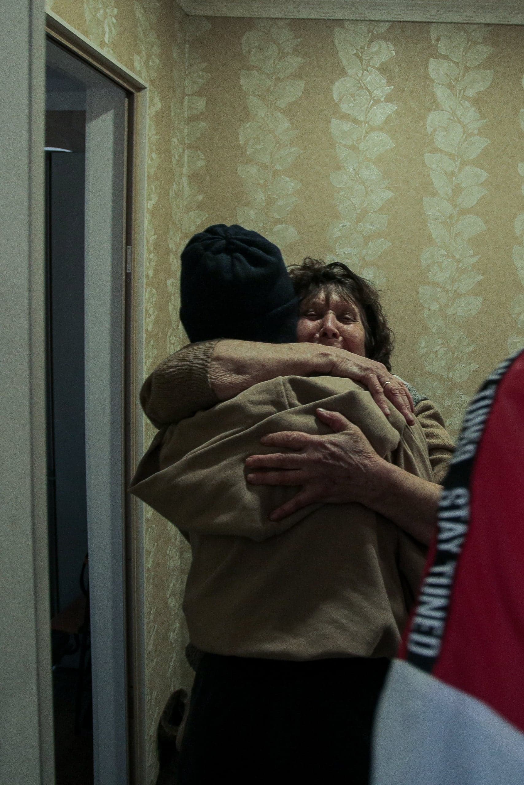 Женщина с закрытыми глазами обнимает человека в бежевом худи и черной шапке. На фоне бежевые обои с узором. Справа виден фрагмент красной одежды с текстом "STAY TUNED".