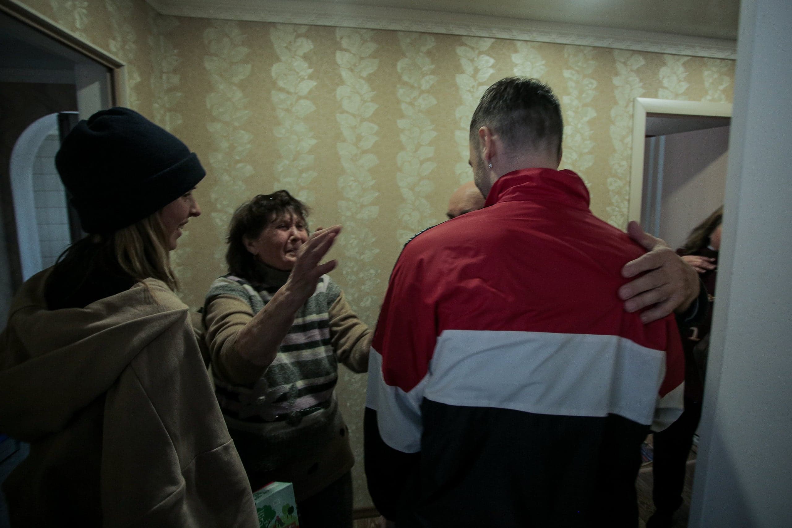 Пожилая женщина, жестикулируя, разговаривает с мужчиной в красно-белой куртке в помещении.