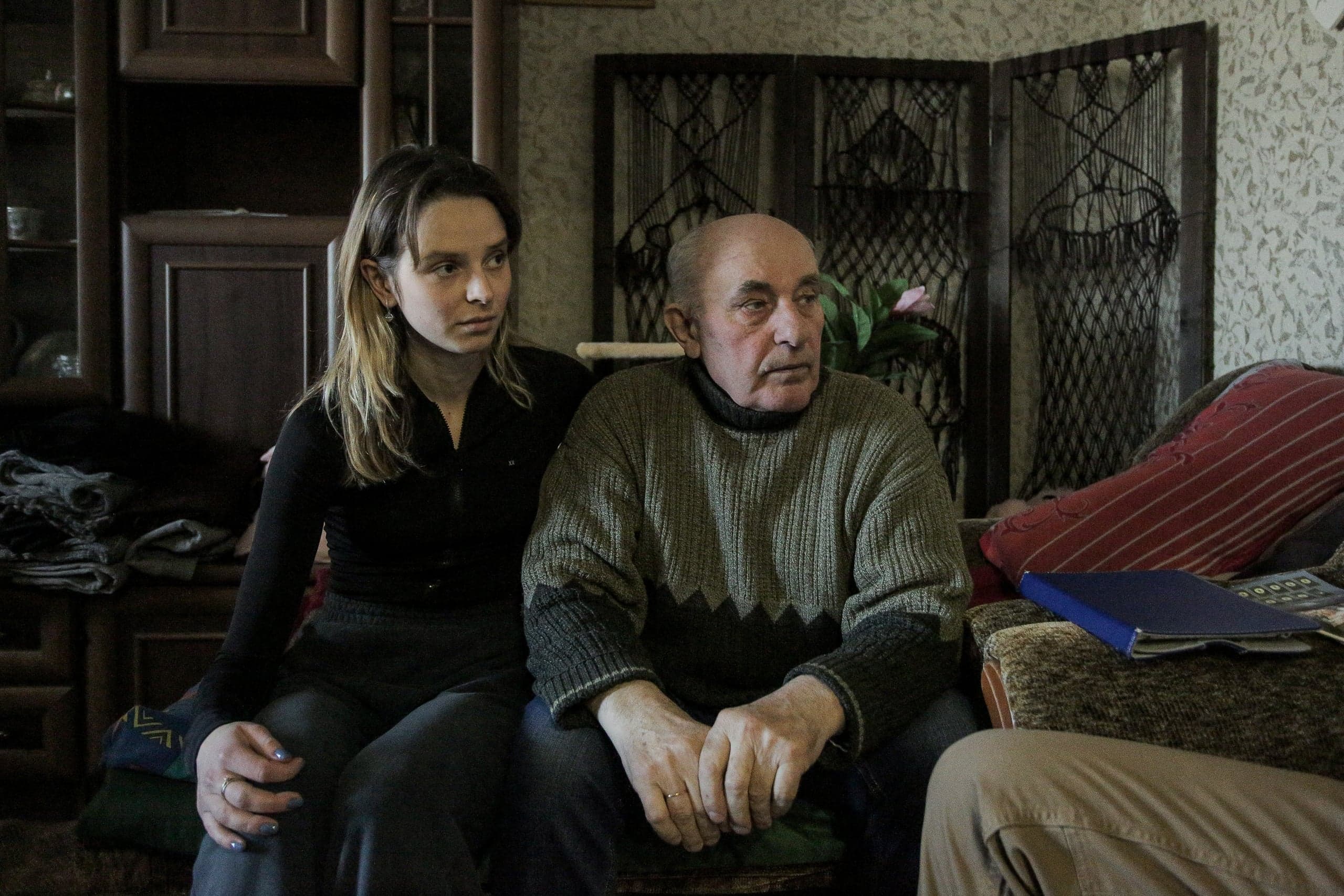 Молодая женщина и пожилой мужчина сидят в тускло освещенной комнате с узорчатыми обоями и старой мебелью.