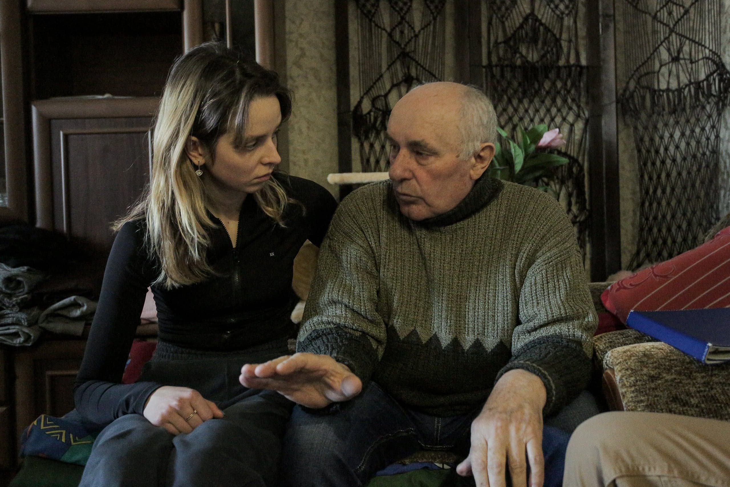 Пожилой мужчина в свитере говорит и жестикулирует, молодая женщина в черной кофте внимательно слушает.