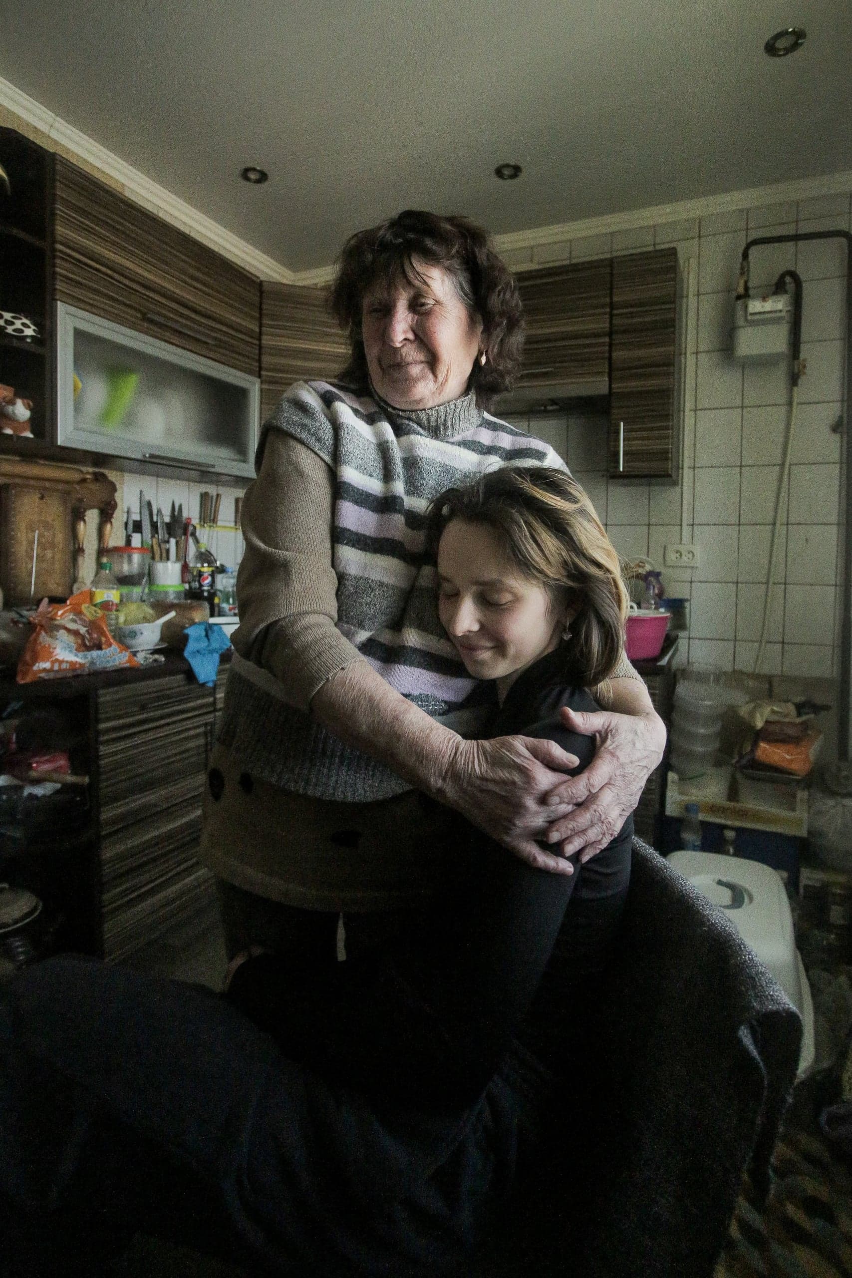 Пожилая женщина обнимает молодую девушку в тесной, заставленной кухонной обстановке.