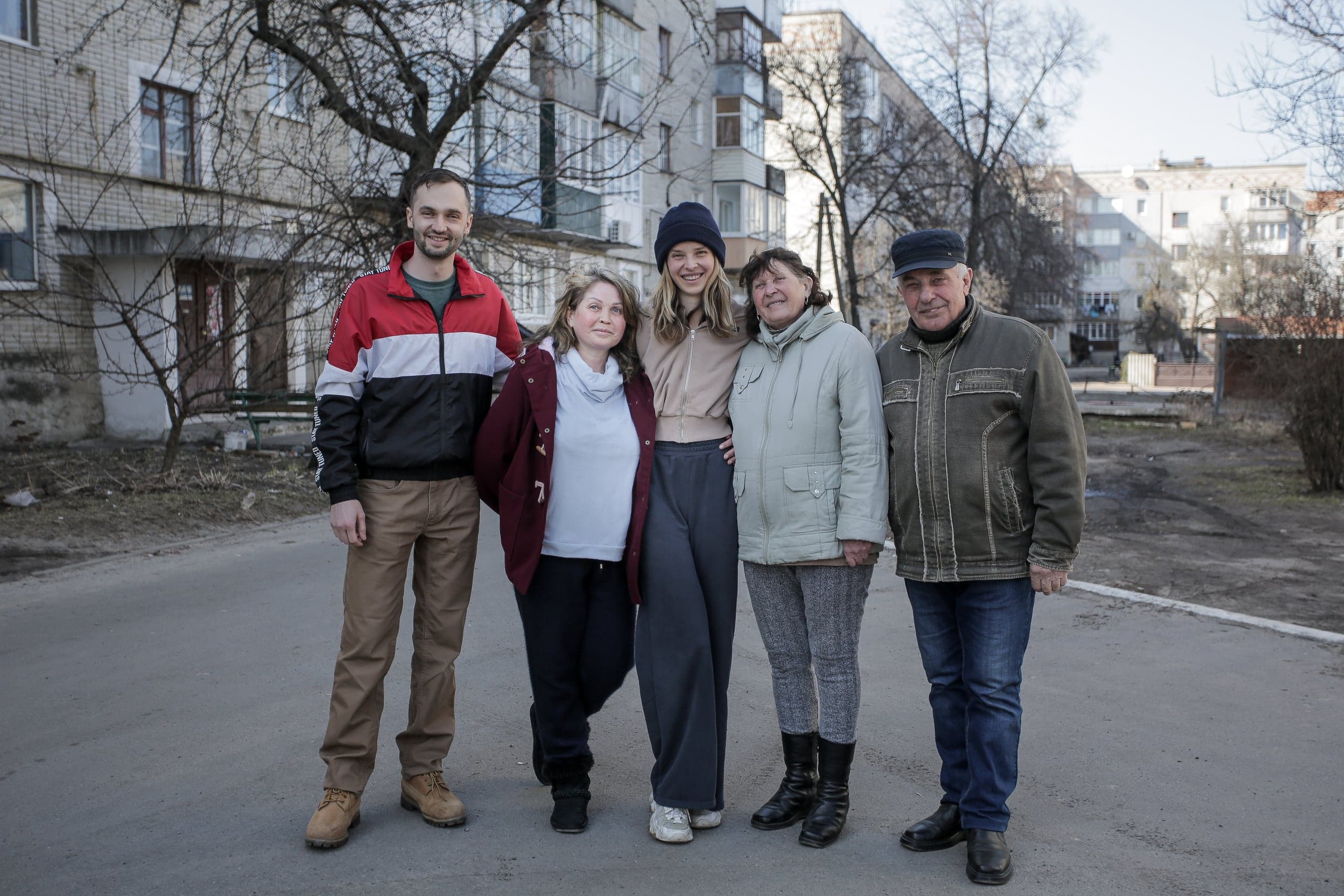 Группа из пяти улыбающихся человек разного возраста (двое мужчин, трое женщин) стоит на улице на фоне жилого дома.