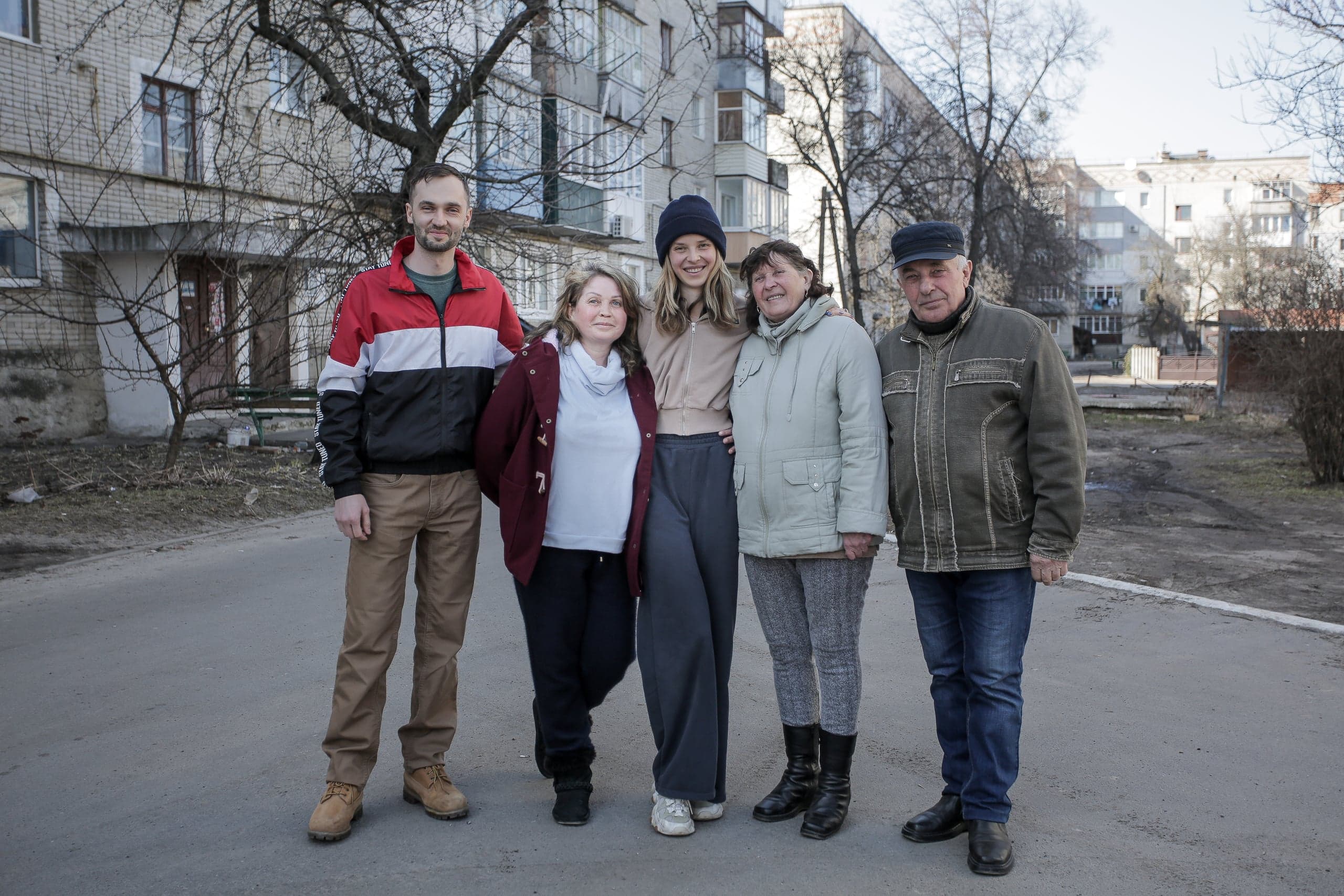 Пять человек (три женщины и двое мужчин разного возраста) стоят на асфальте во дворе жилого квартала.