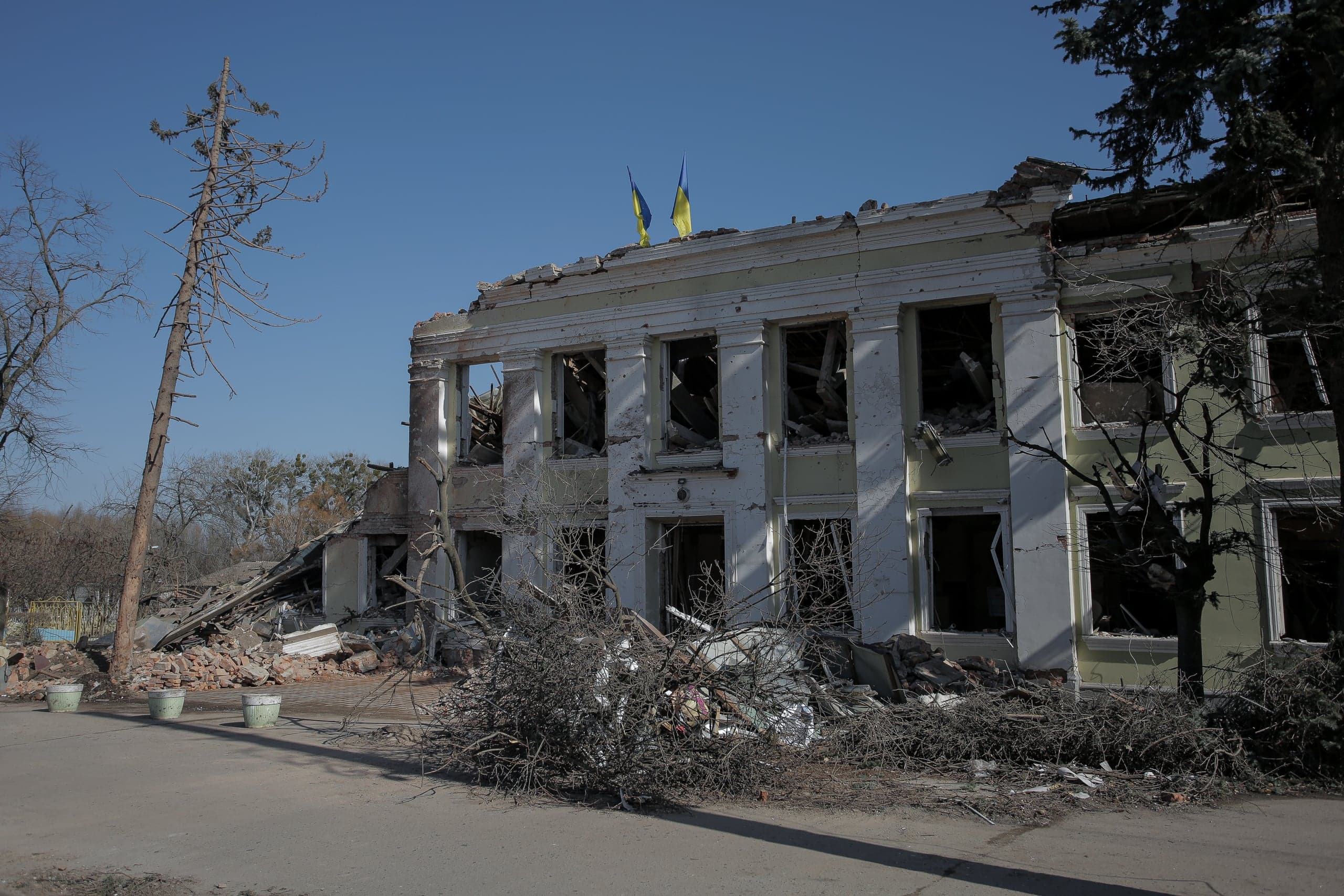Разрушенное двухэтажное здание с выбитыми окнами и двумя флагами Украины на крыше, вокруг обломки и ветки.