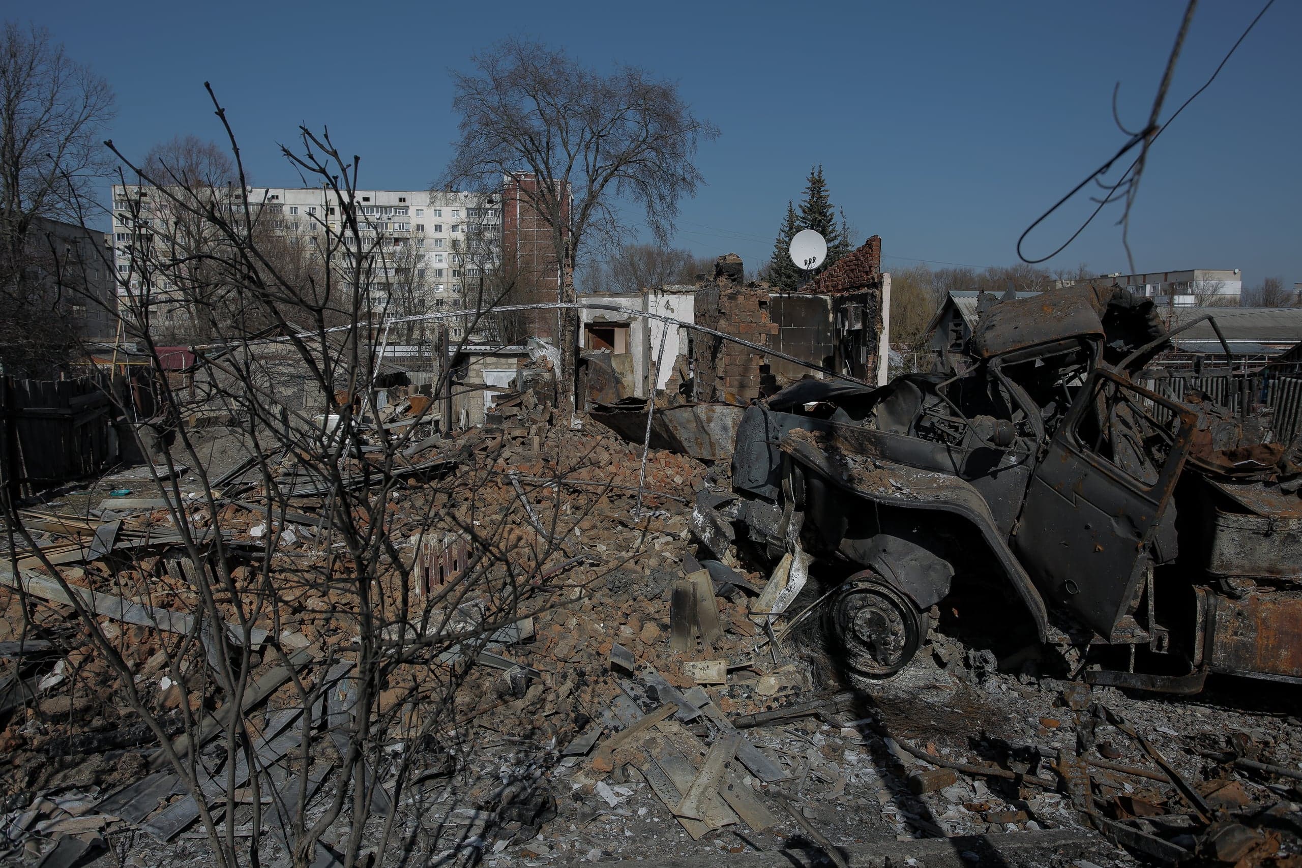 Разрушенная территория с обломками домов и сгоревшим автомобилем на фоне уцелевшего многоэтажного здания.