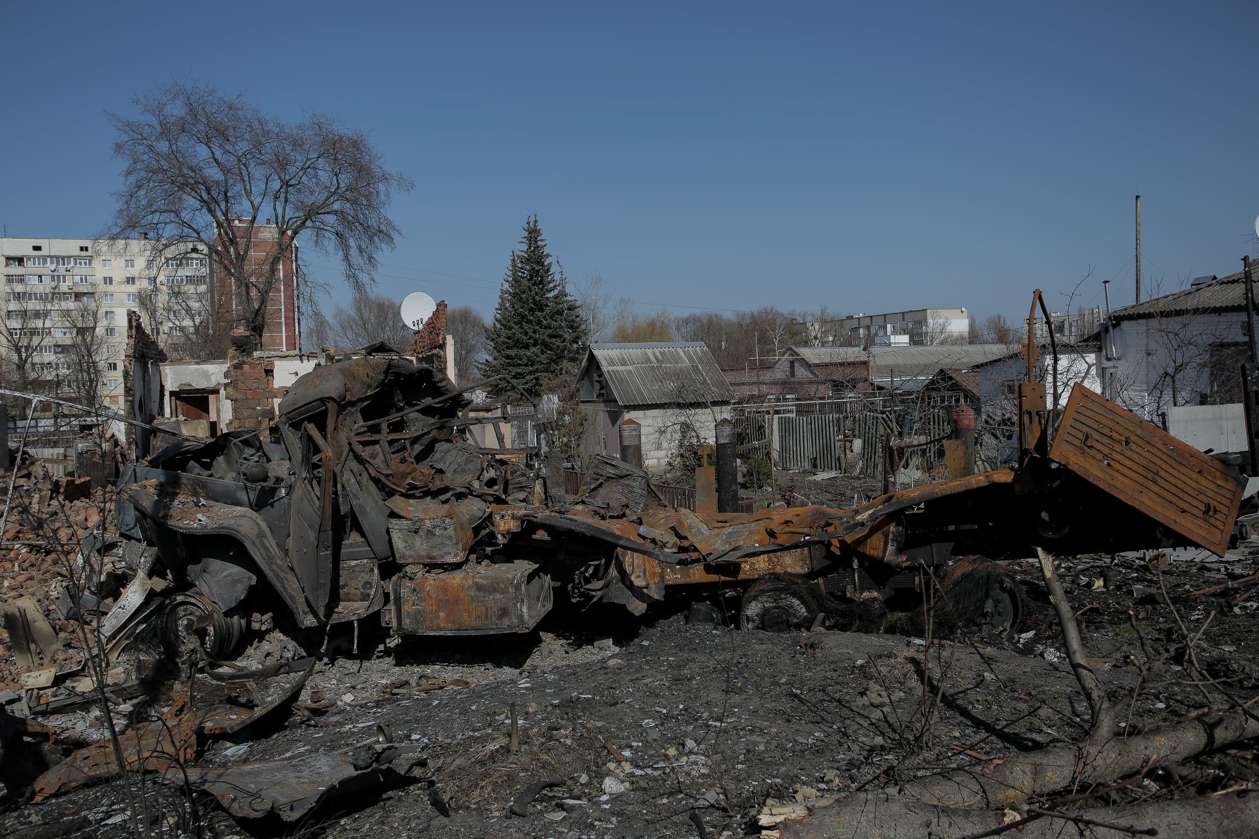 Сгоревшие остатки военной техники на фоне разрушенных домов частного сектора и многоэтажных зданий.