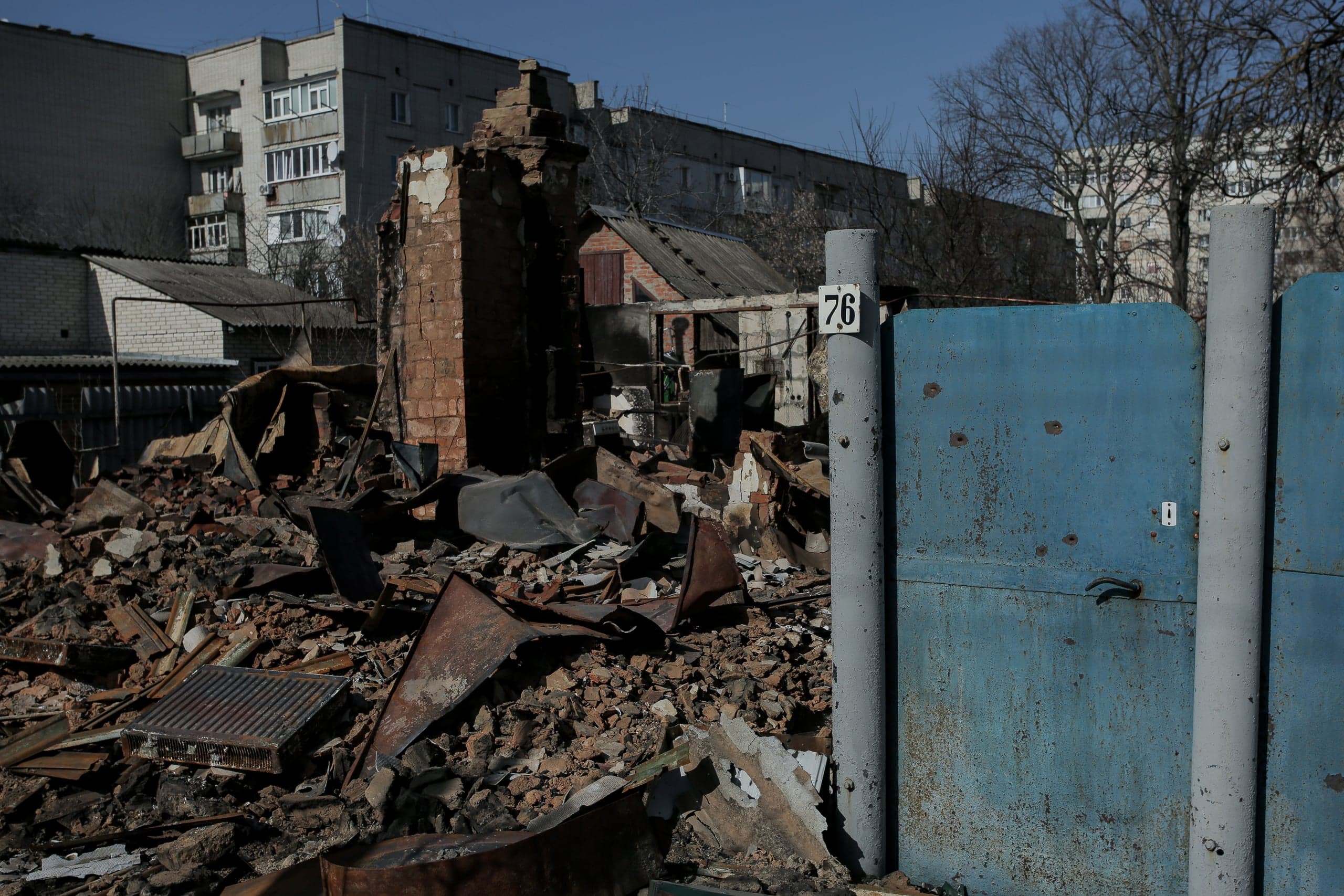 Руины частного дома с грудой обломков и частичной кирпичной стеной. Справа — синие металлические ворота с номером "76". На заднем плане — жилой многоэтажный дом.