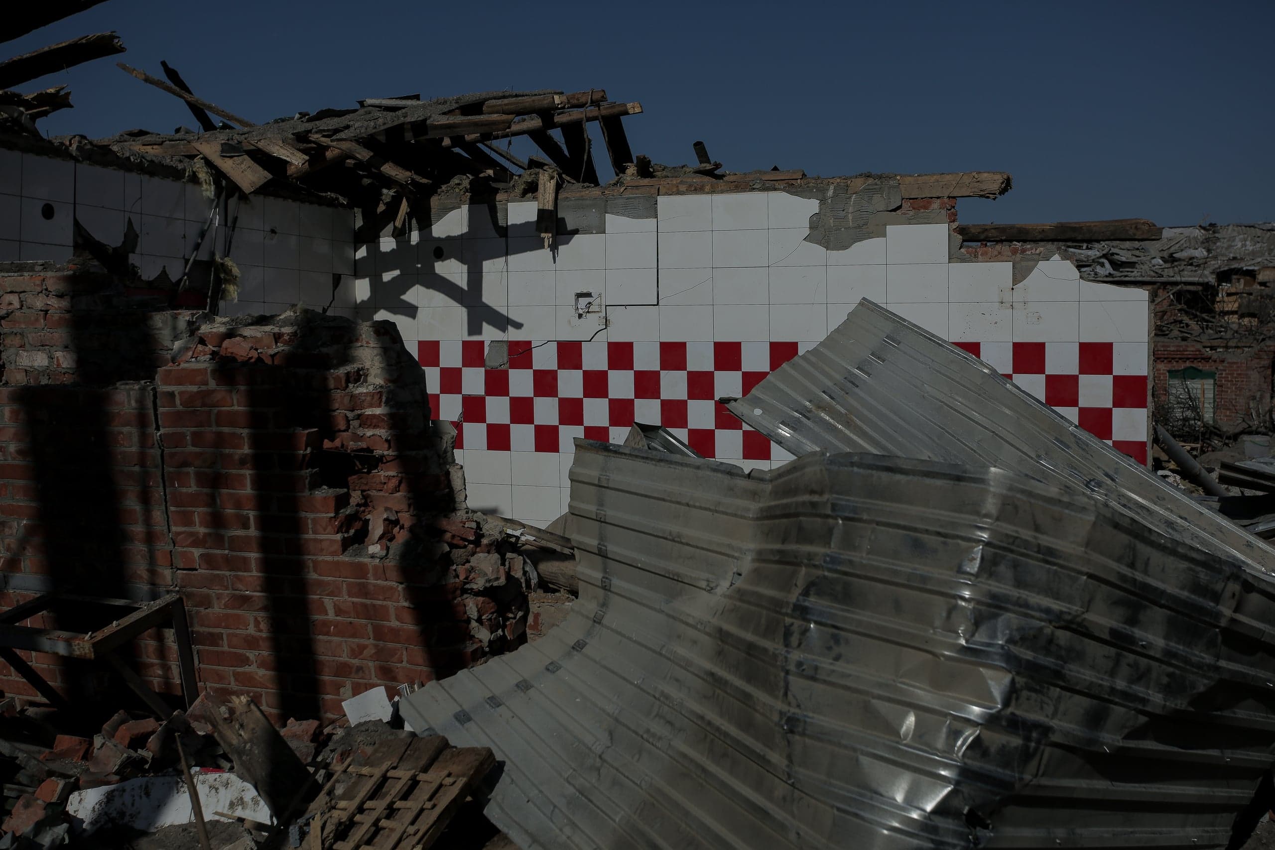 Разрушенное здание: уцелевший фрагмент стены с красно-белой клетчатой плиткой среди кирпичных обломков и смятого гофрированного металла.
