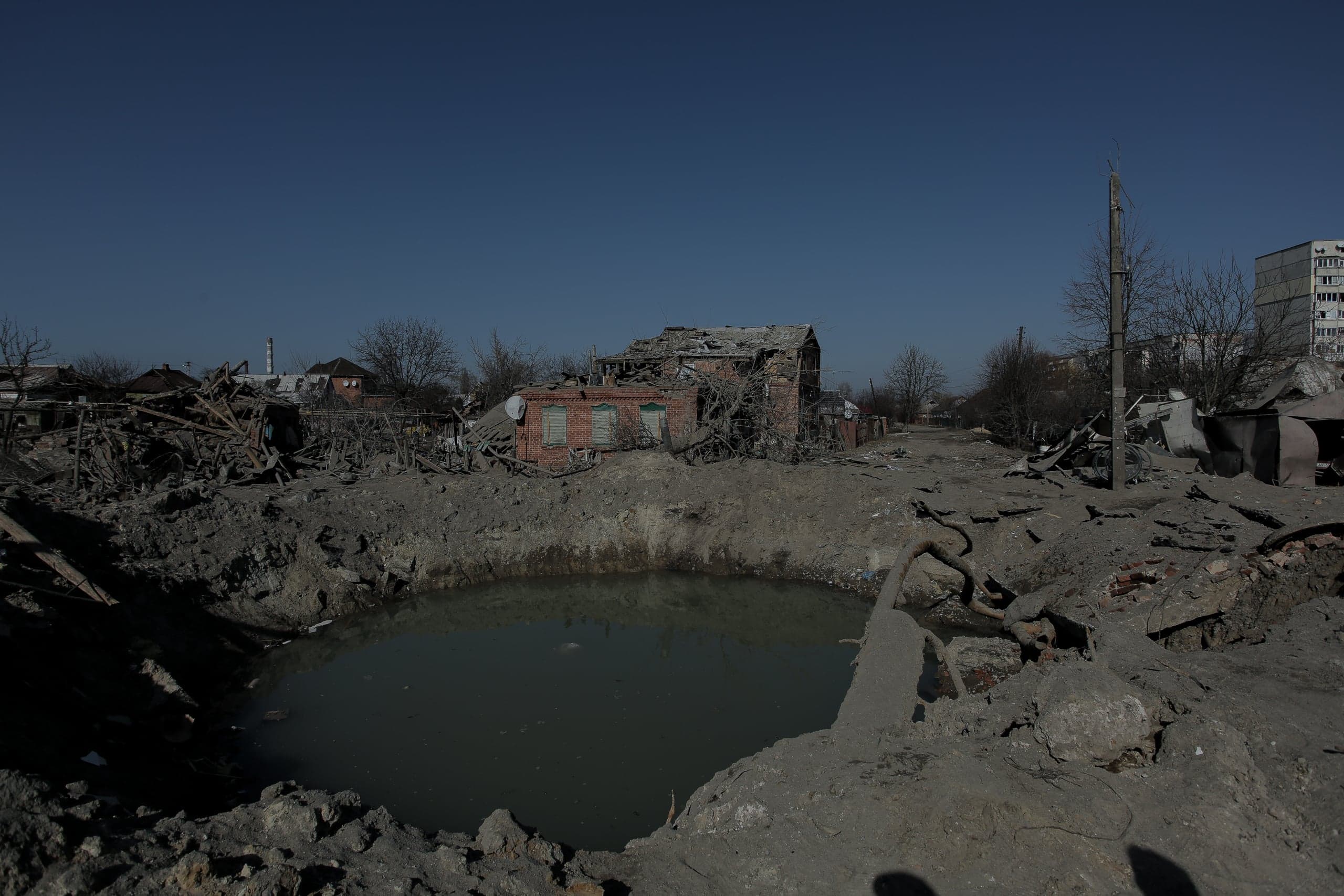 Большая воронка, заполненная водой, посреди разрушенного жилого квартала. На заднем плане — руины кирпичных и панельных домов.