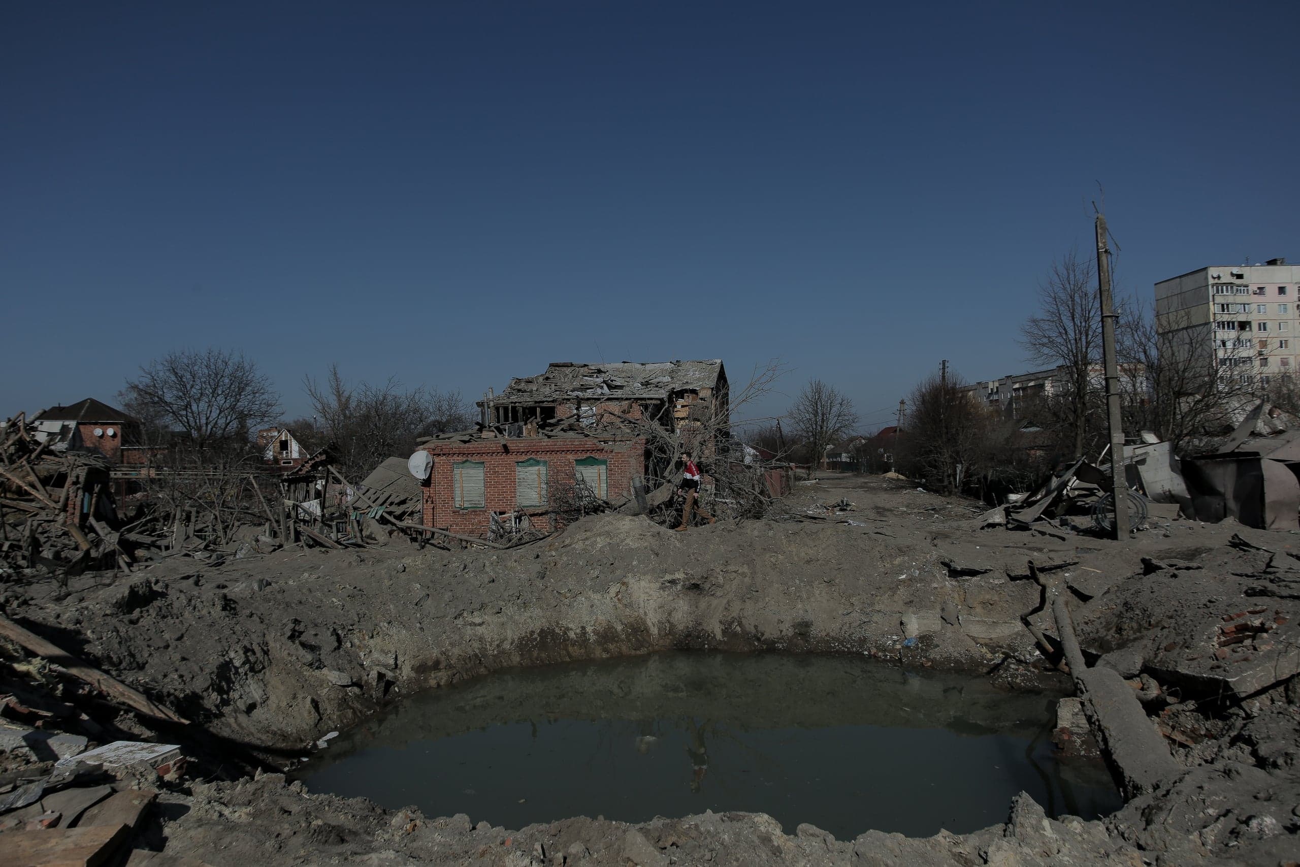 Воронка с водой на переднем плане; за ней разрушенный кирпичный дом, обломки и уцелевшие многоэтажки на горизонте.