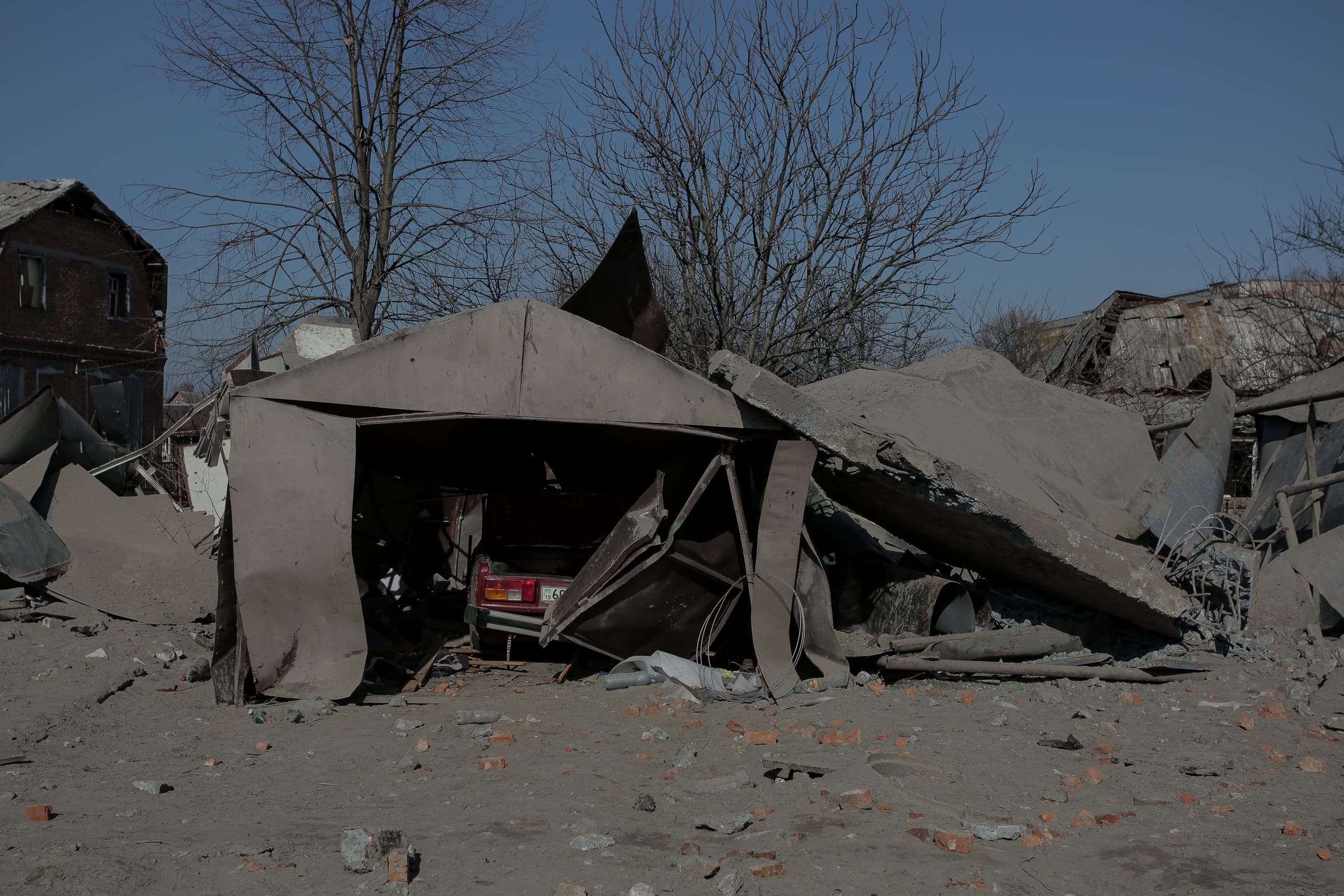 Разрушенный гаражом с обломками и красным автомобилем внутри. На заднем плане руины домов и сухие деревья.