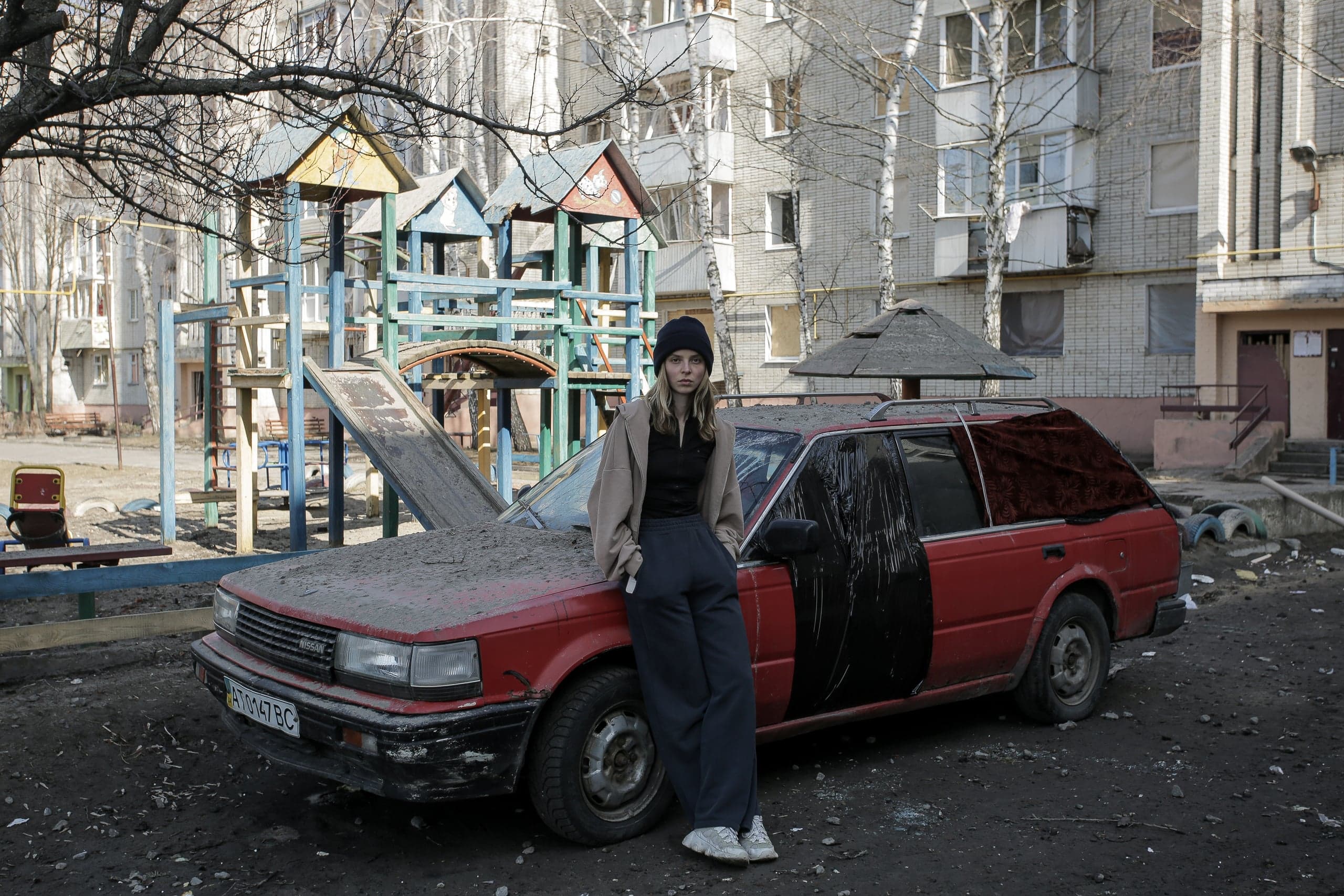 Женщина опирается на покрытый пылью красный универсал с номером "AT 0447 BC" на фоне разрушенной детской площадки и жилого дома.