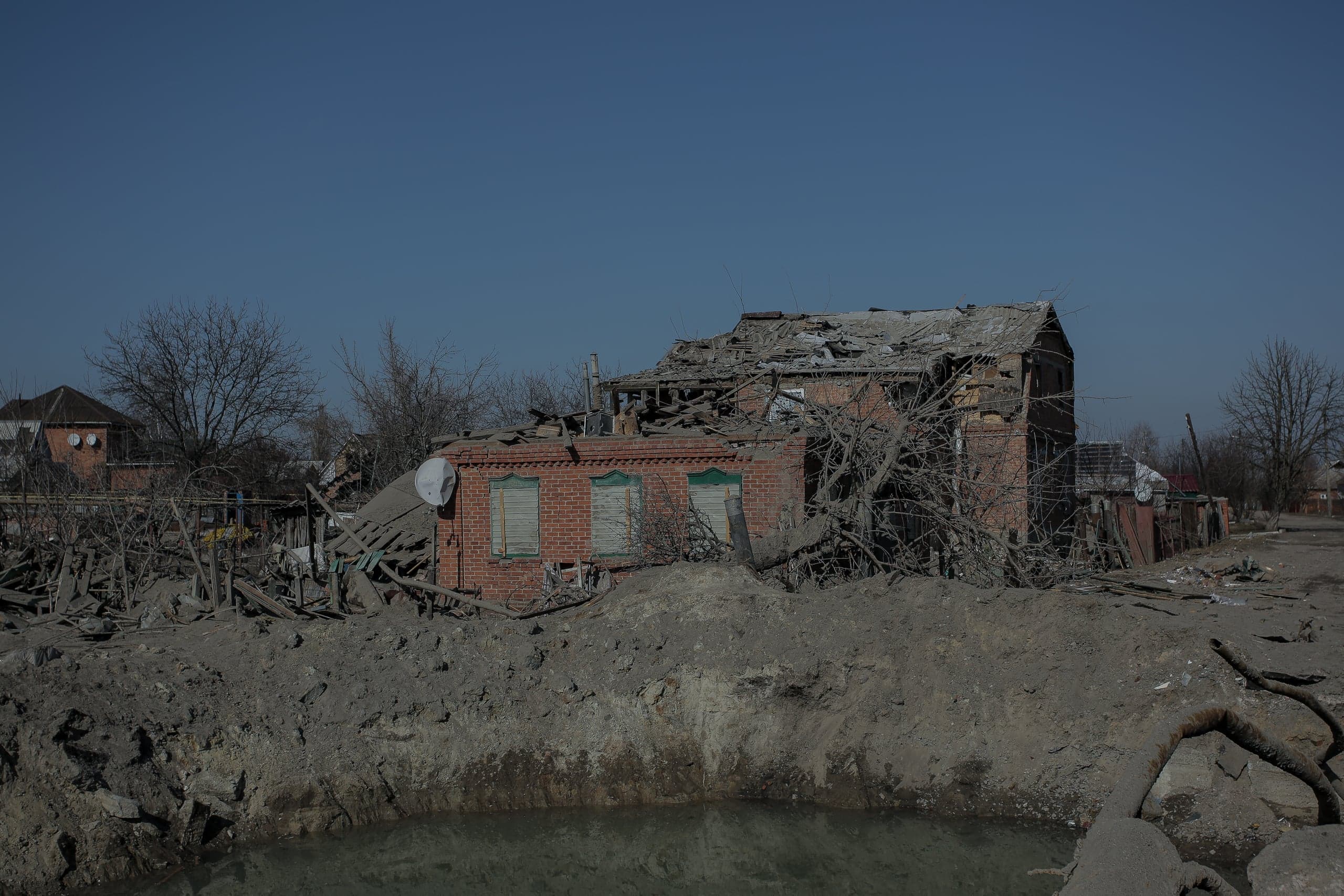 Разрушенный кирпичный дом и большая воронка от взрыва, заполненная водой, на переднем плане.