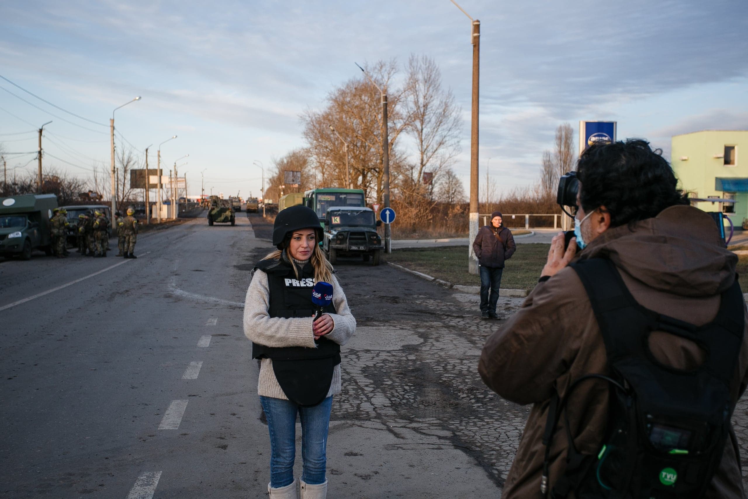 Журналист в каске и бронежилете с надписью "PRESS" ведет репортаж на дороге, позади видны военные грузовики и солдаты. Оператор снимает ее.