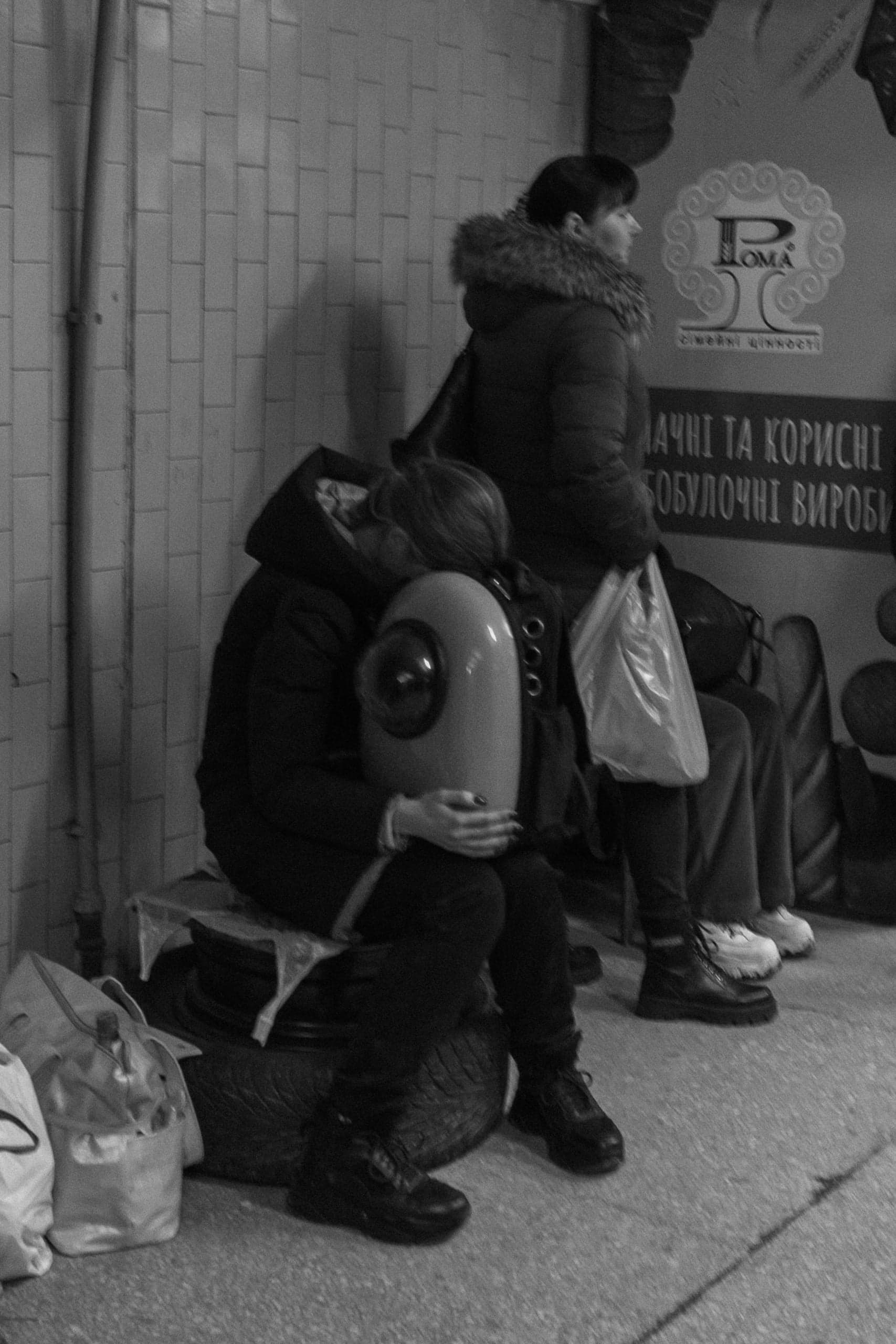 Чёрно-белое фото двух женщин, одна из которых обнимает футуристическую переноску для животного. На стене надписи: "P LOMA", "сімейні цінності" и "ВДАЧНІ ТА КОРИСНІ ЯБЛУЧНІ ВИРОБИ".