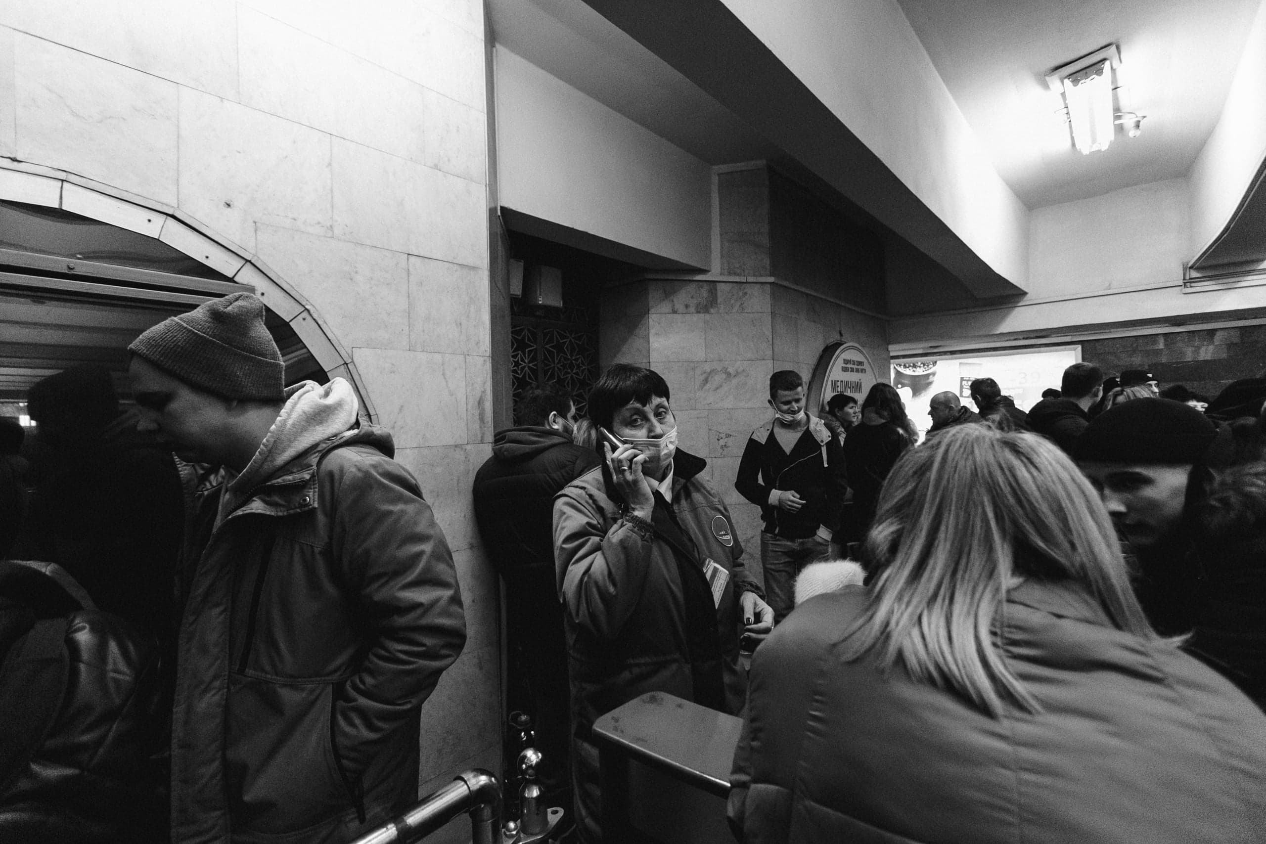 Чёрно-белое фото толпы в вестибюле. Женщина в маске разговаривает по телефону. Виден знак "ВЕЖЛИВЫЙ".