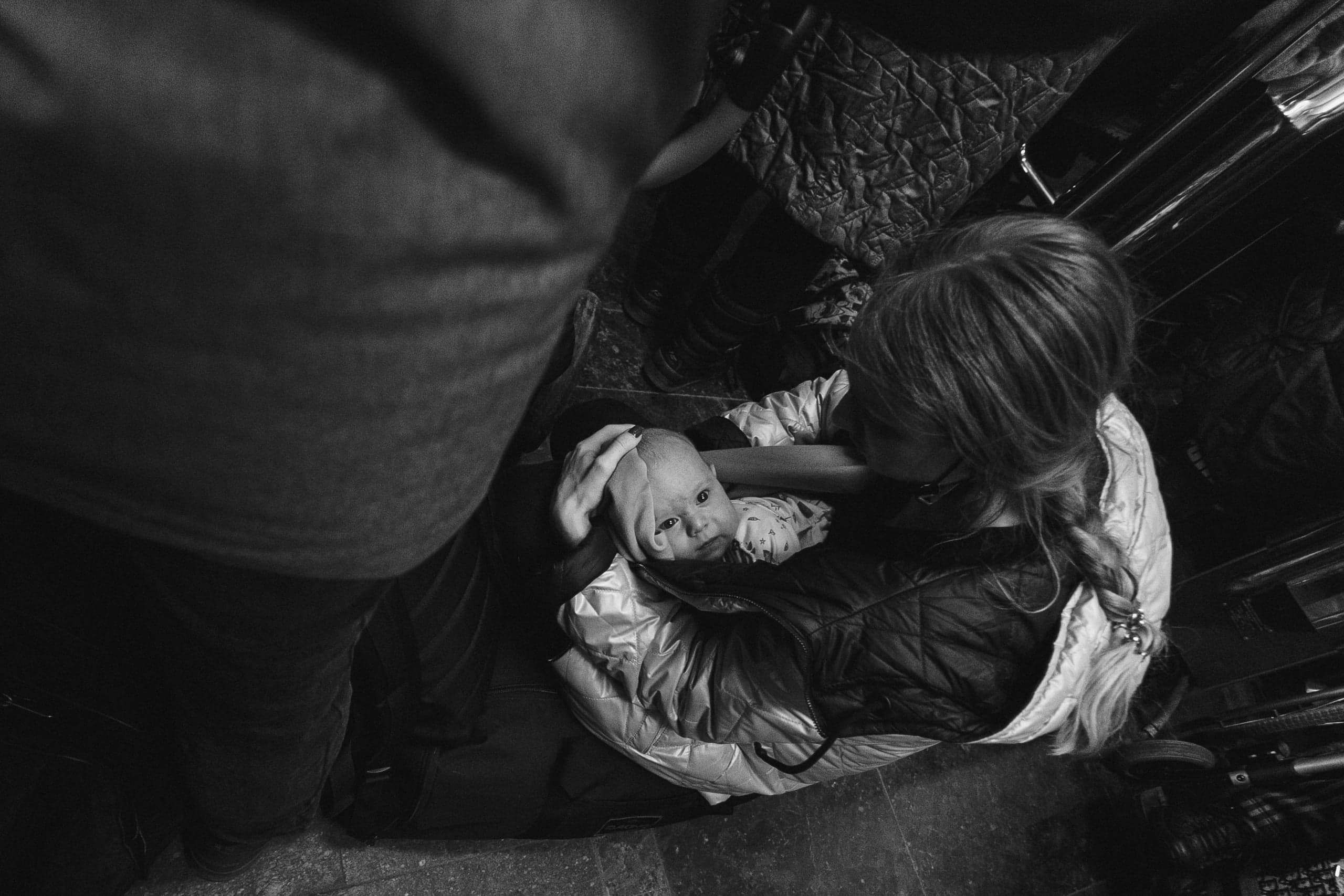 Черно-белое фото, снятое сверху: Женщина в куртке держит младенца, нежно гладя его по голове. Слева видна часть фигуры стоящего человека.