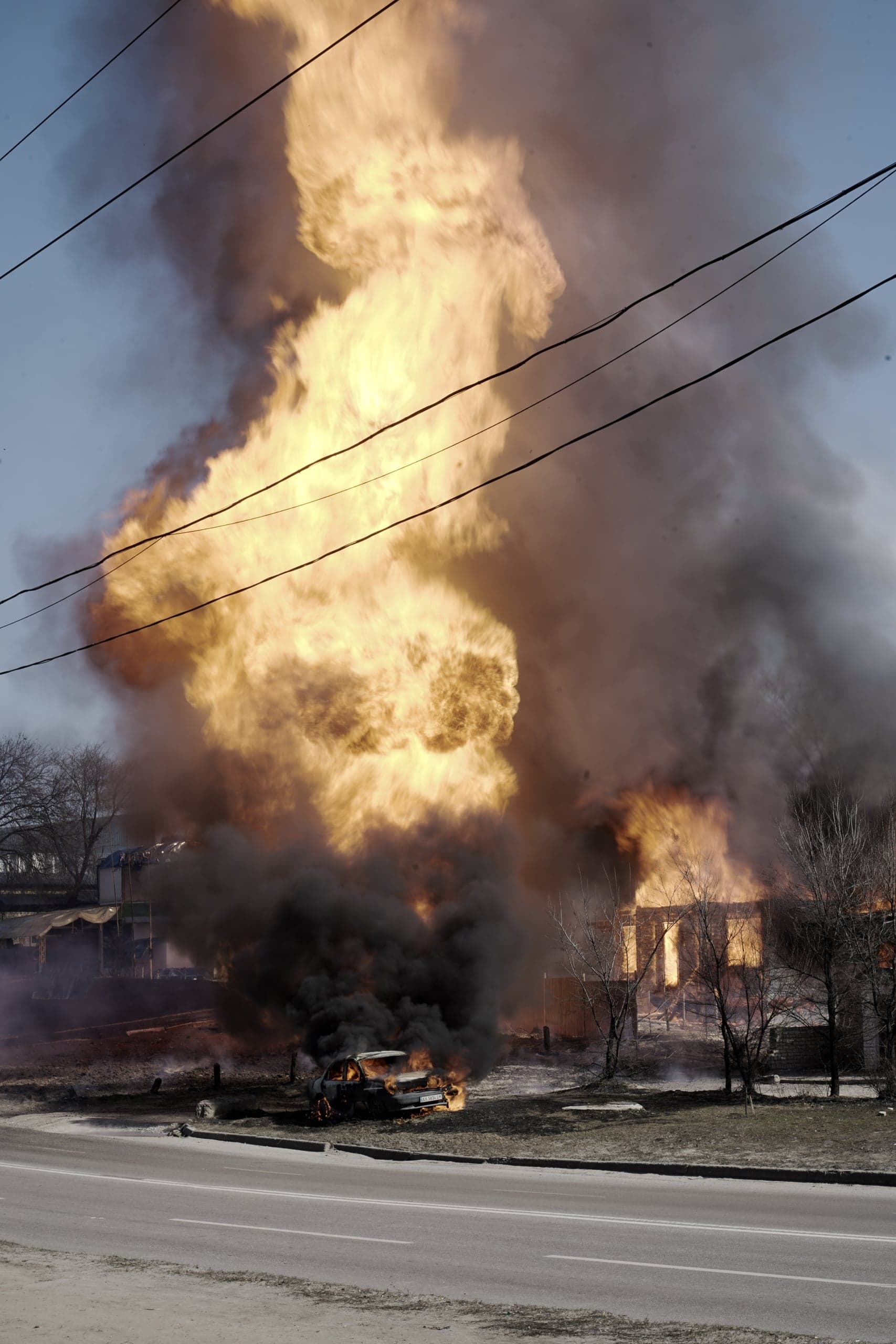 Огромное облако яркого пламени и черного дыма поднимается над горящим зданием, рядом с которым у обочины дороги стоит объятый огнем разрушенный автомобиль.