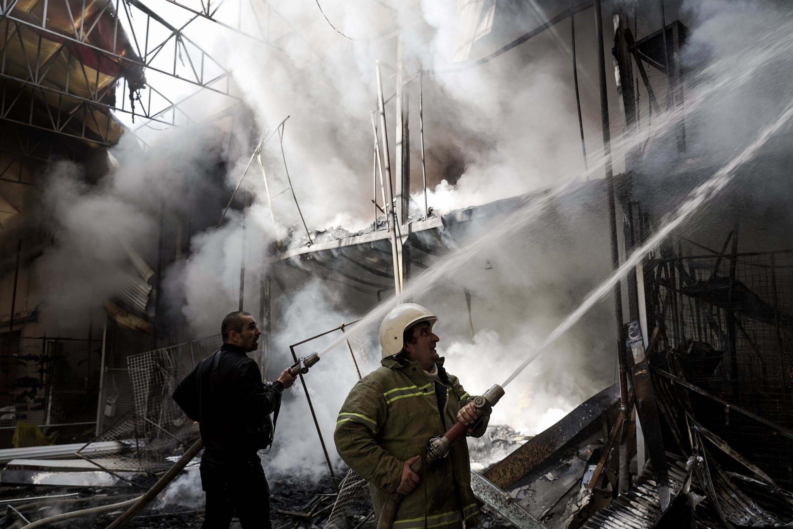 Два пожарных направляют мощные струи воды в задымленное, обгоревшее помещение разрушенного здания. Один пожарный в защитной форме и каске, другой — в темной одежде.