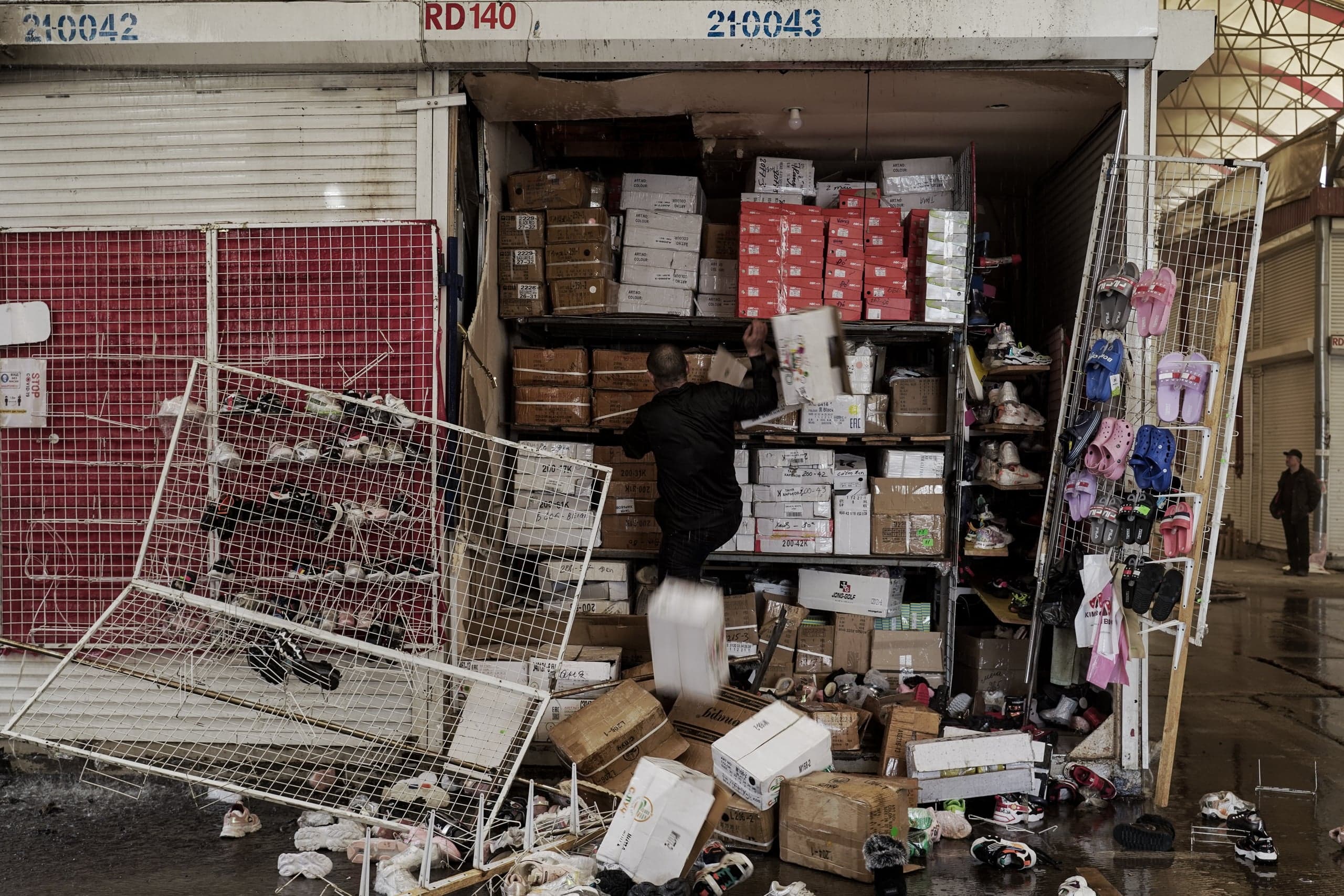 Мужчина в черном спешно заталкивает коробки с обувью в переполненный рыночный киоск. На мокром полу перед киоском валяются упавшие металлические стеллажи и разбросанная обувь.