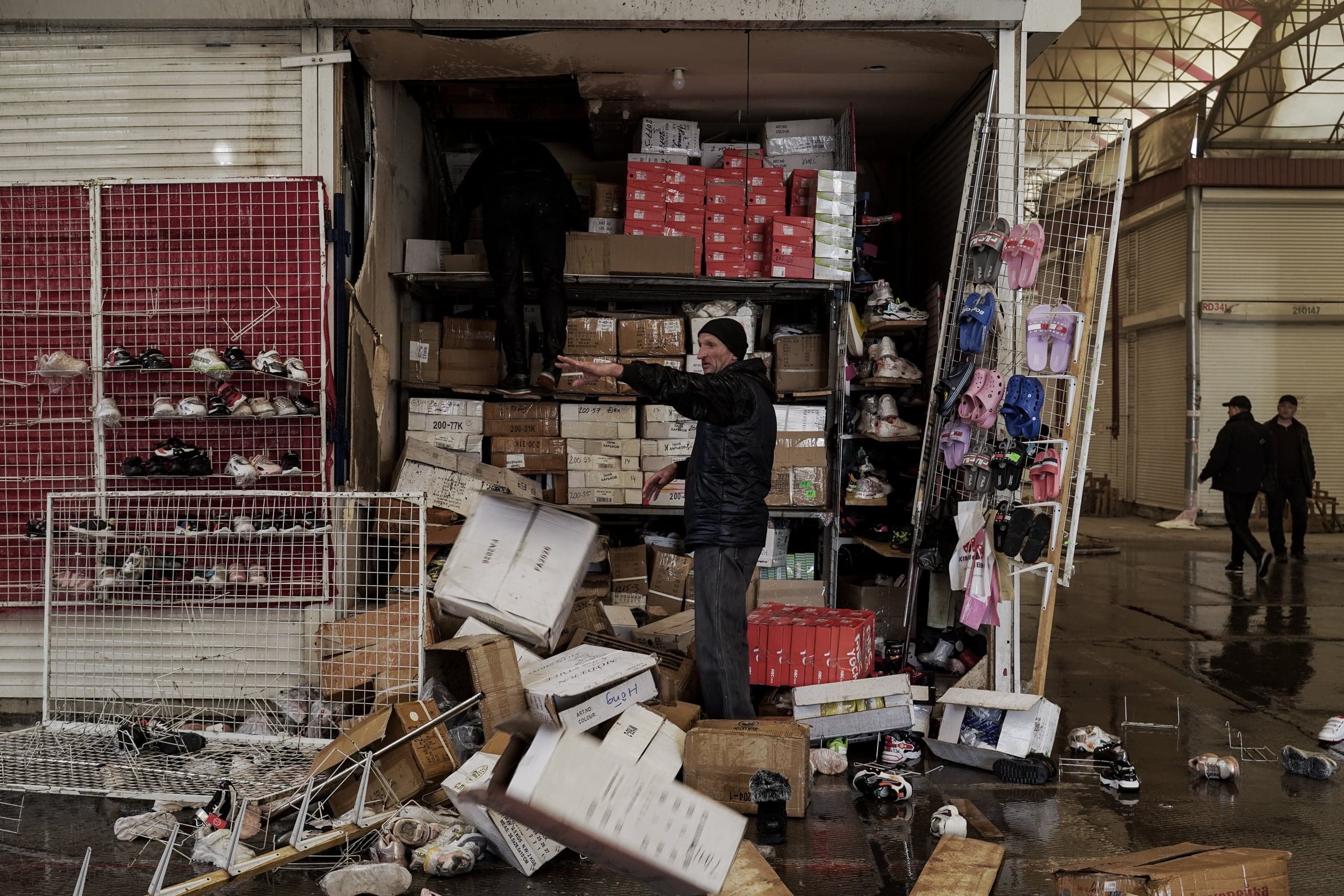 Мужчина стоит посреди груды коробок и обуви перед переполненным ларьком на мокром полу рынка; второй человек виден на верхних полках.