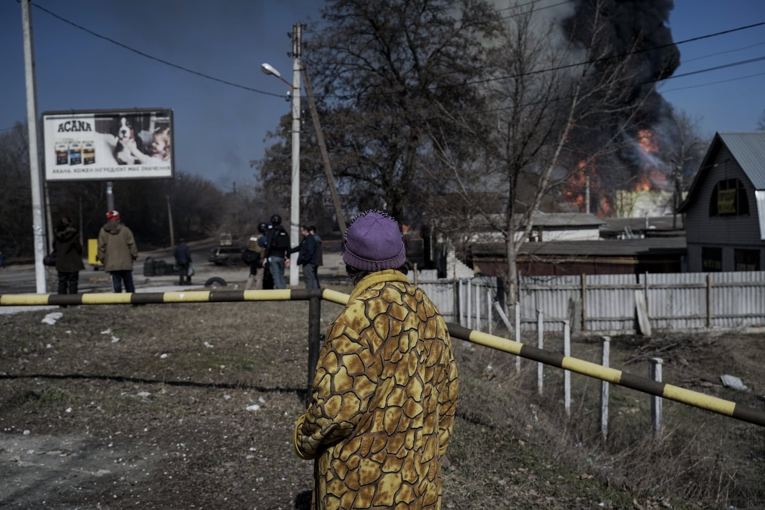 Пожилая женщина в желтом пальто и фиолетовой шапке стоит спиной к зрителю за шлагбаумом, наблюдая за сильным пожаром и густым черным дымом, поднимающимся над жилыми домами вдали. На заднем плане также видны журналисты и рекламный щит.