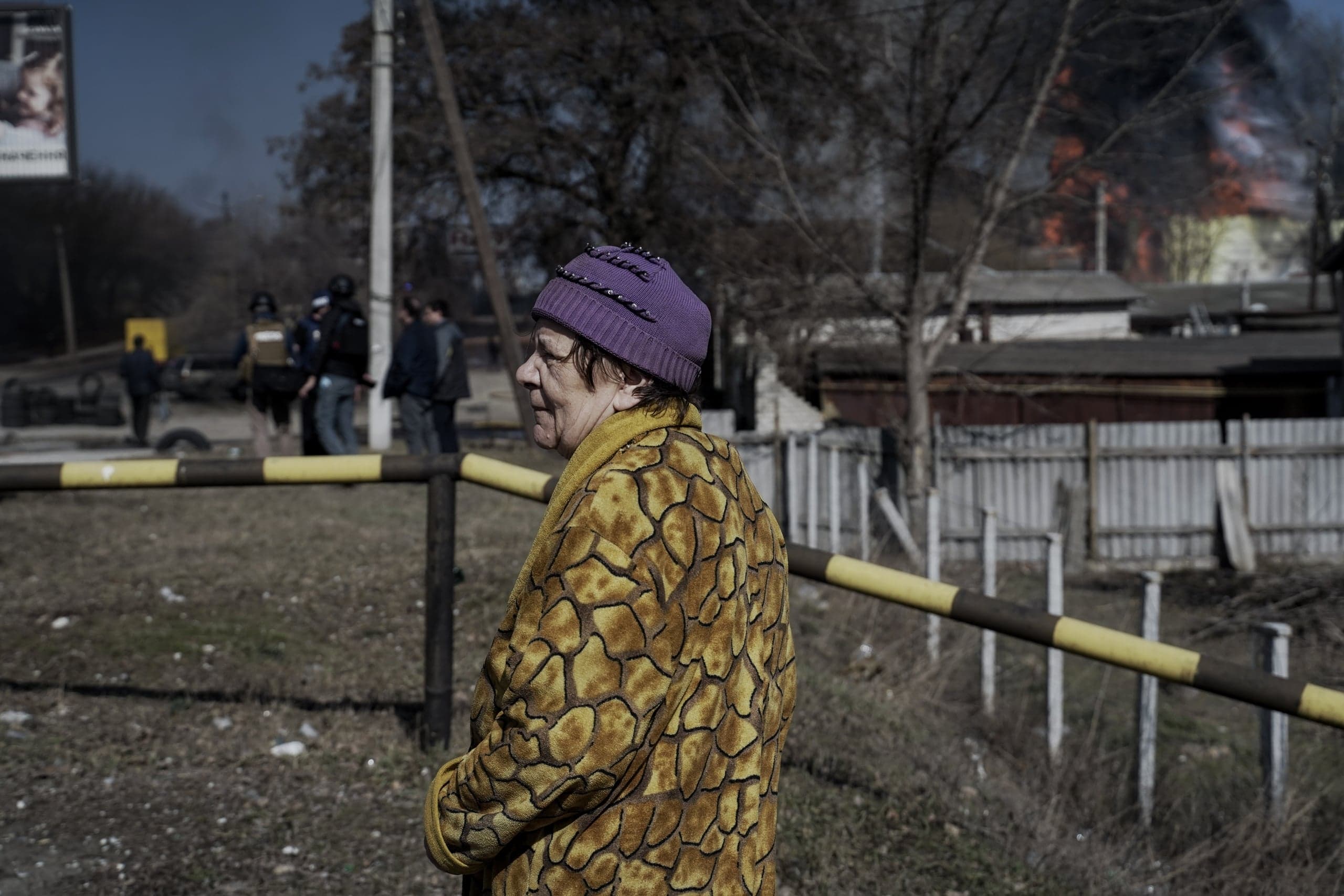 Пожилая женщина в фиолетовой шапке и желтой узорчатой куртке стоит у шлагбаума. На заднем плане видно сильное пламя и черный дым от пожара.