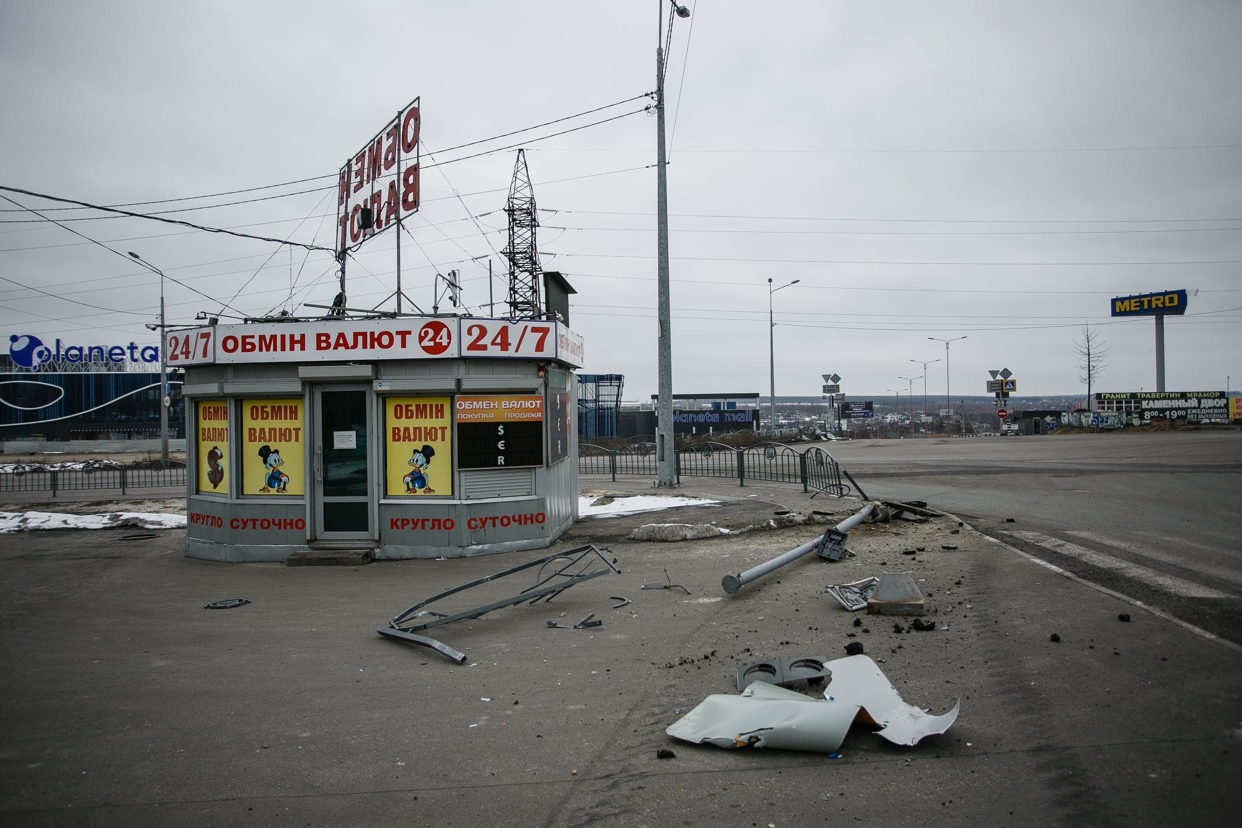 Обменный пункт ("ОБМІН ВАЛЮТ 24/7") на улице. На асфальте перед киоском лежат обломки и сломанный столб. На заднем плане видны вывески "Planeta" и "METRO".