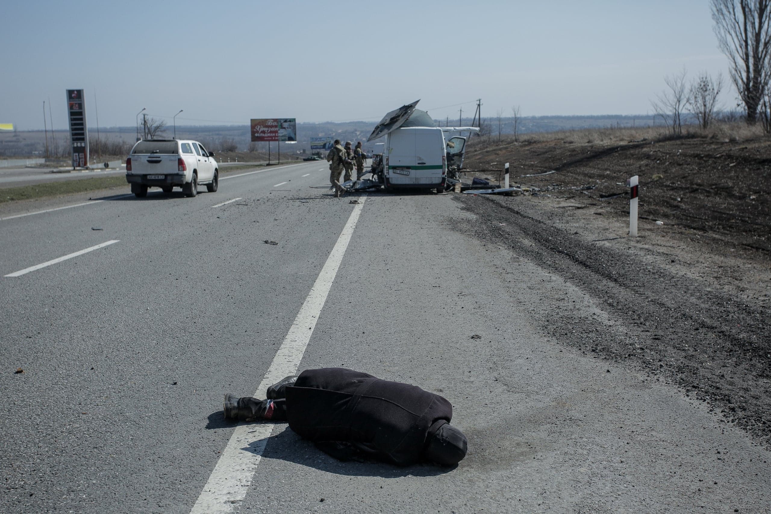 Тело лежит на шоссе перед поврежденным белым фургоном, вокруг которого стоят военнослужащие. Дорога усыпана обломками.
