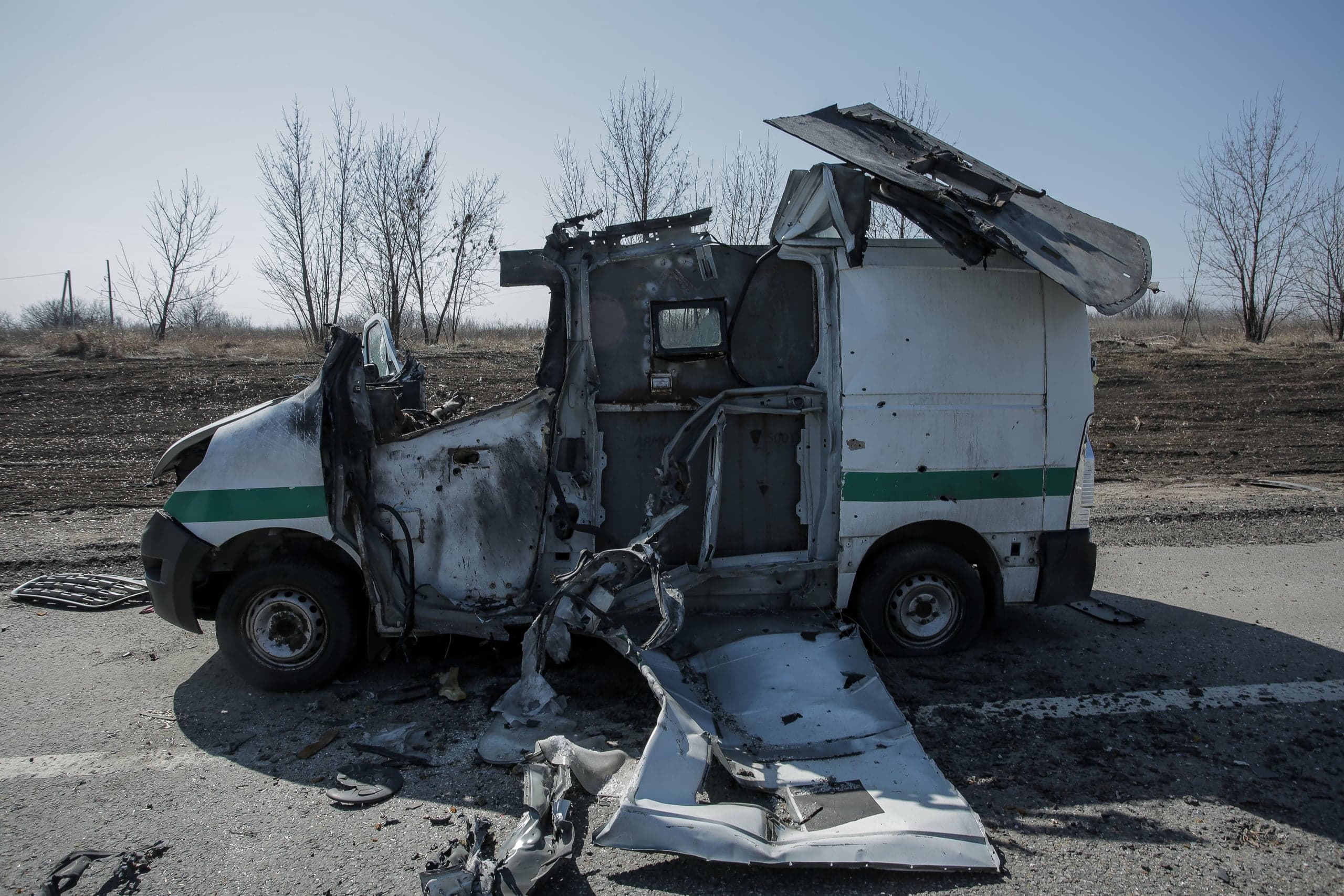 Сожжённый и сильно повреждённый белый бронированный инкассаторский фургон с зелёной полосой, стоящий на обочине дороги.