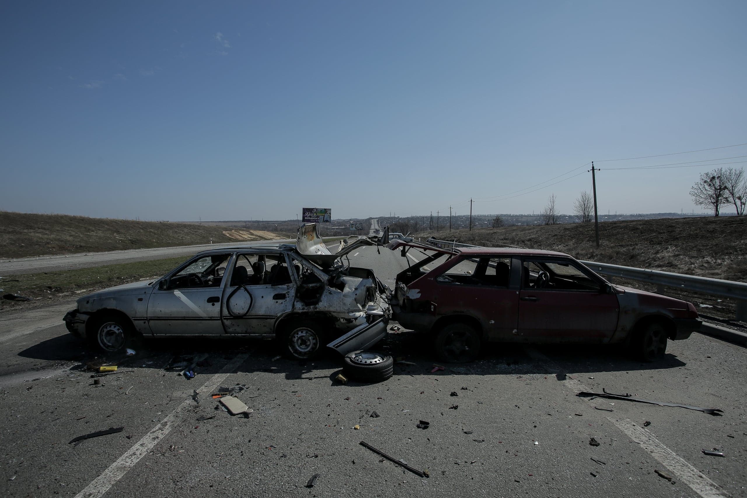 Два разбитых и изрешеченных пулями автомобиля (серебристый и красный) стоят на шоссе, вокруг обломки.
