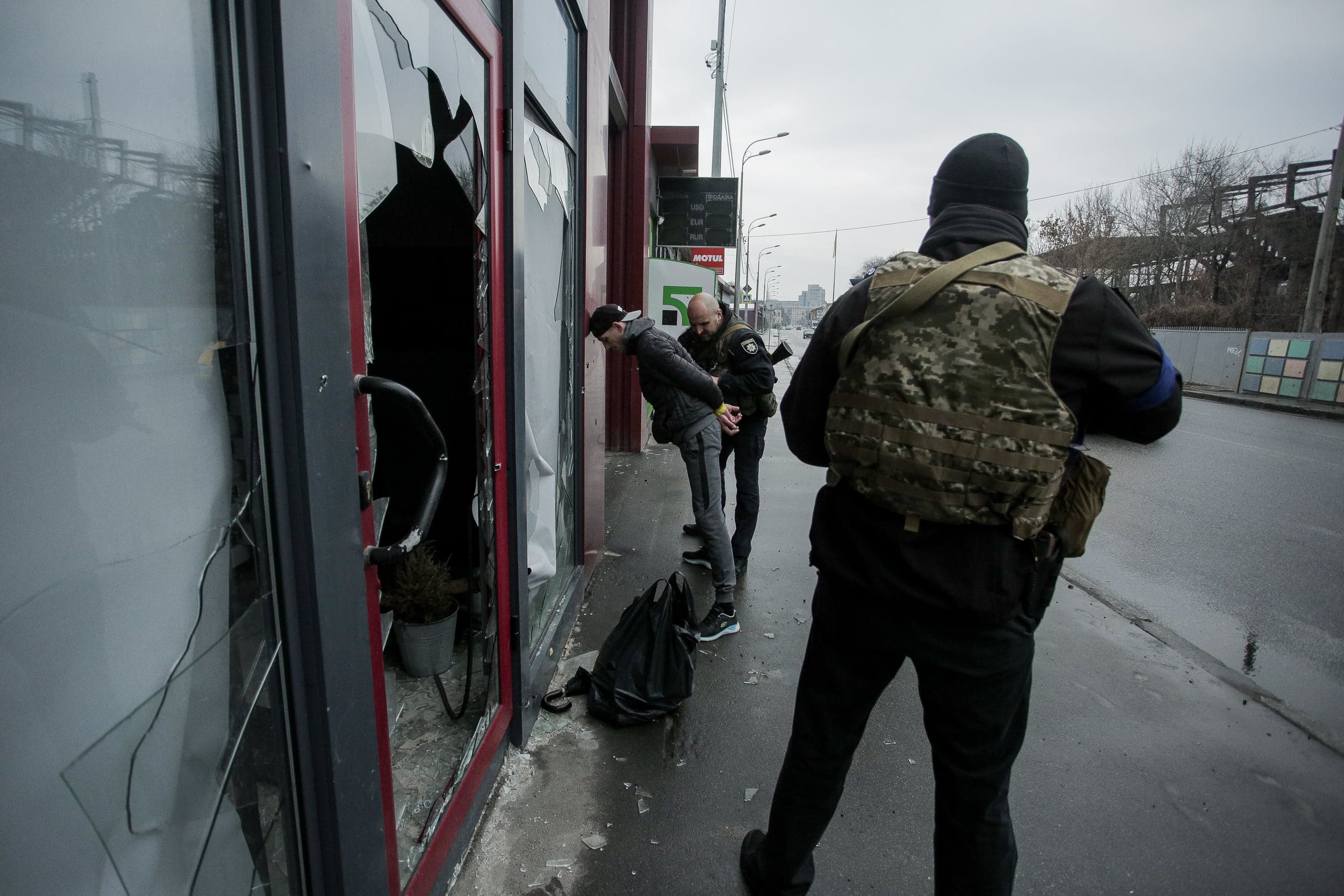 Задержание мужчины сотрудником у разбитой витрины магазина. Вооруженный человек в бронежилете стоит спиной.
