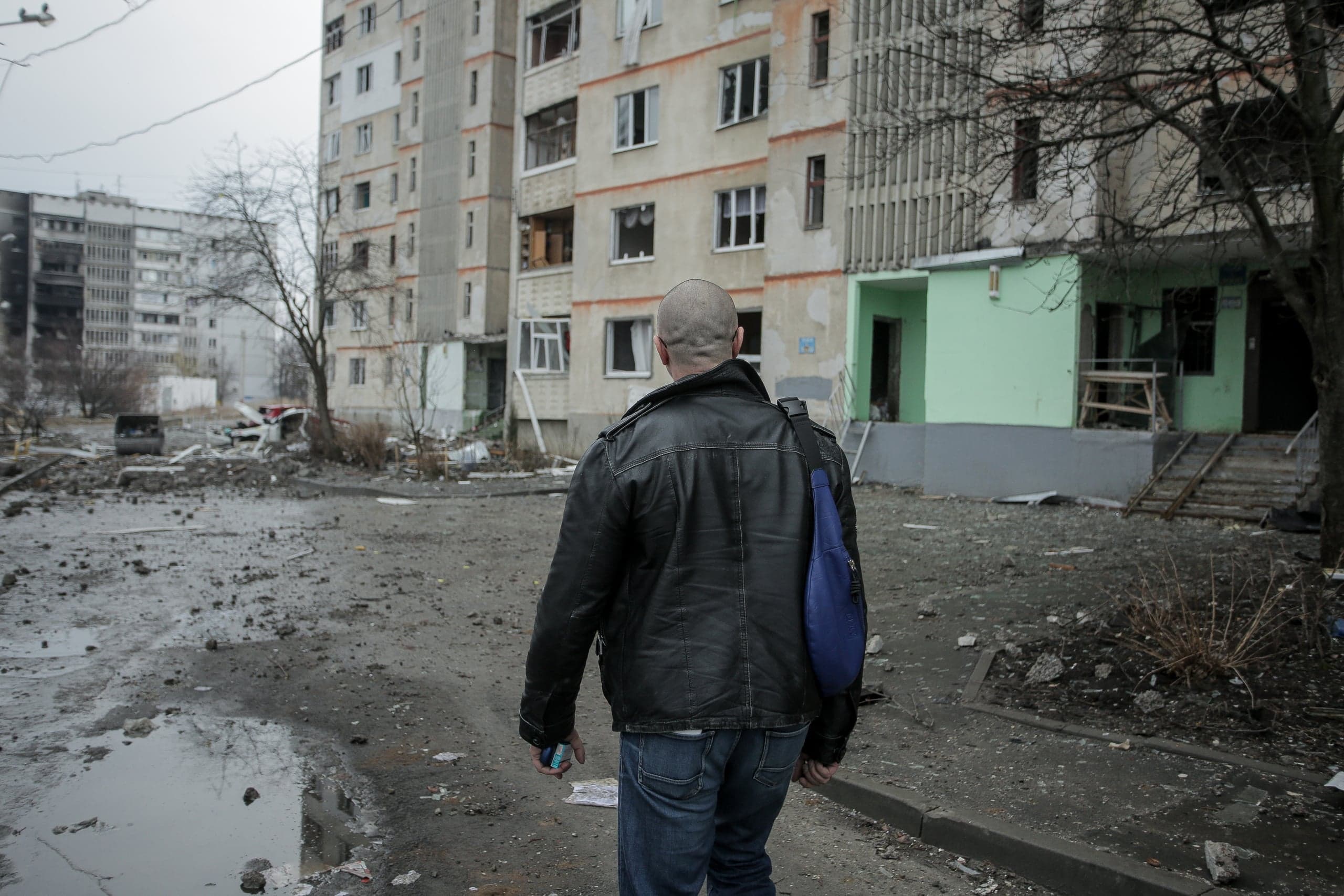 Мужчина в кожаной куртке идет через разрушенный двор мимо поврежденных многоэтажных домов.