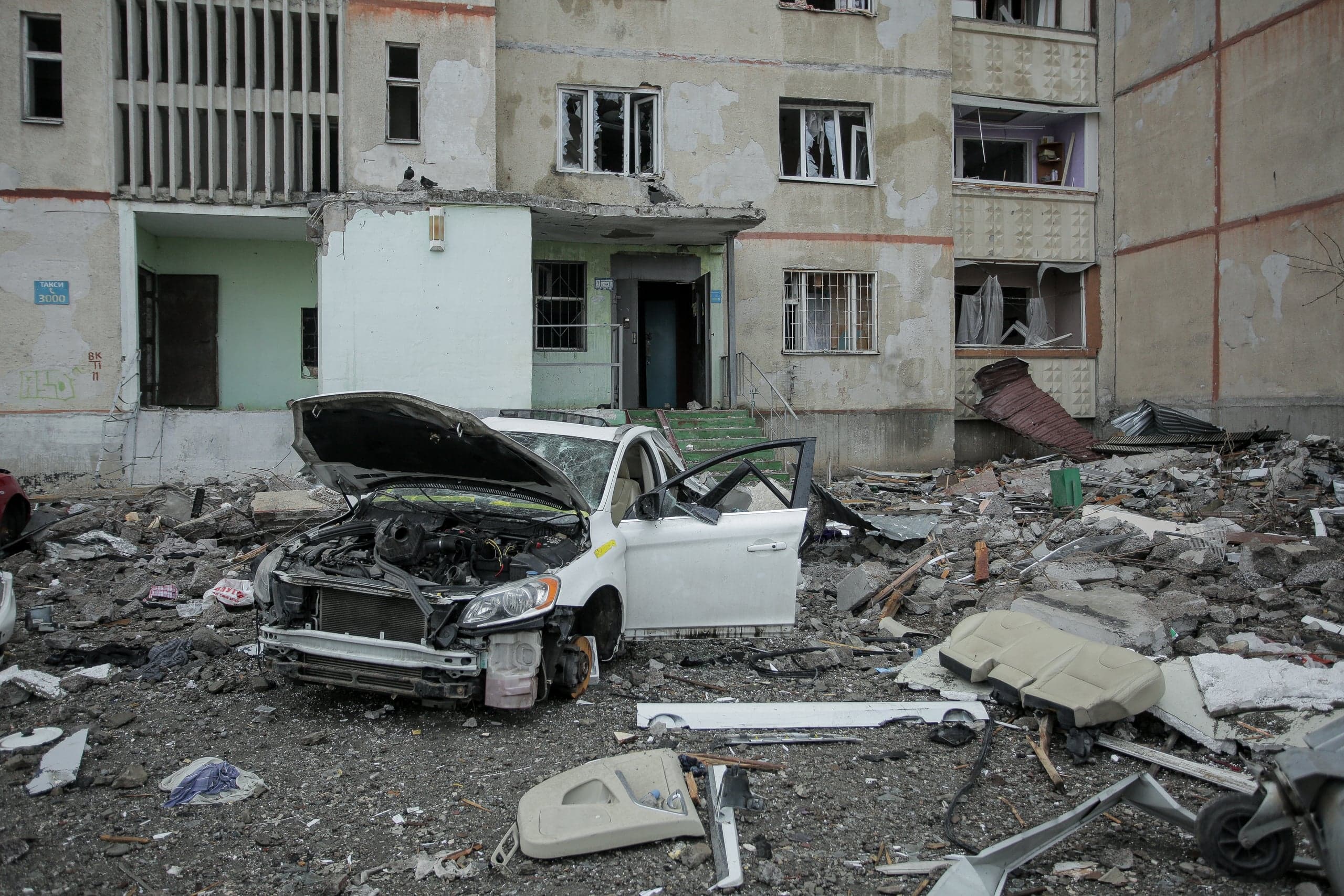 Разрушенный белый автомобиль и груда обломков перед поврежденным многоэтажным домом с выбитыми окнами.