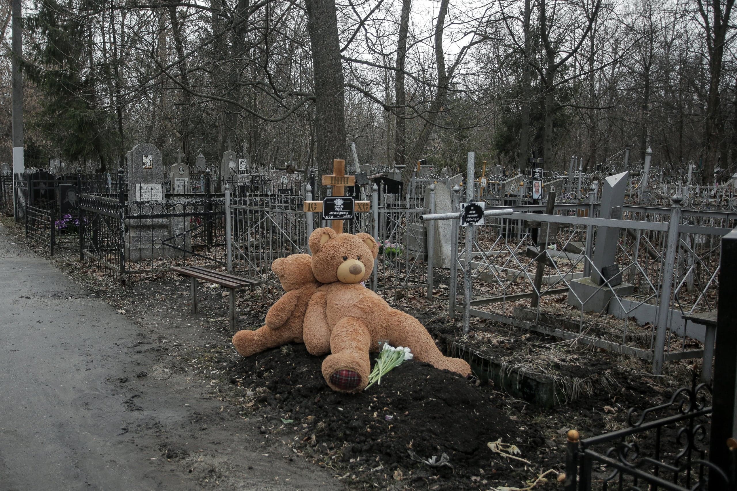 Два плюшевых медведя и белые цветы лежат на свежей могиле на фоне мрачного кладбища.