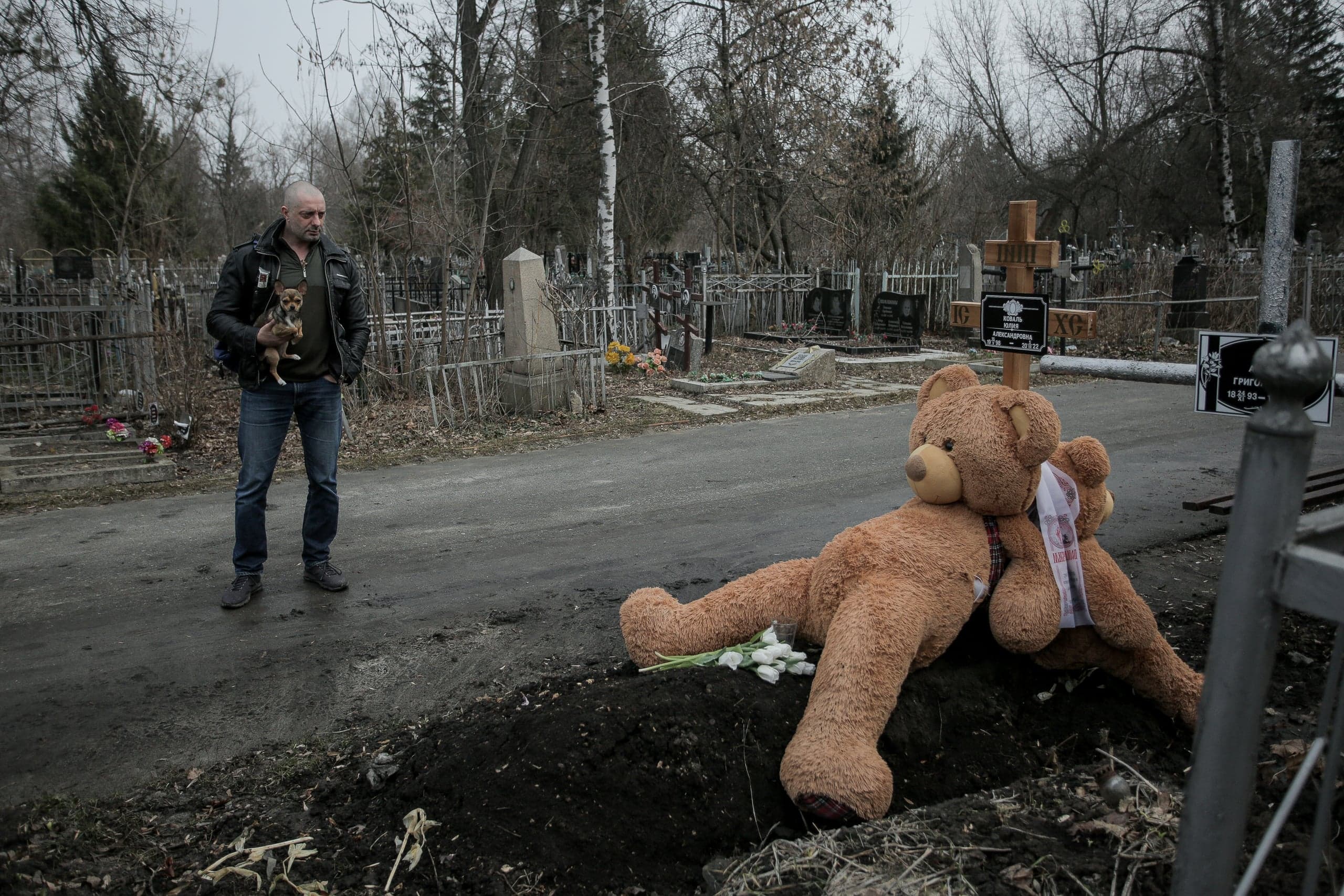 Мужчина с собакой стоит на кладбище; на свежей могиле лежит большой плюшевый медведь и белые тюльпаны.
