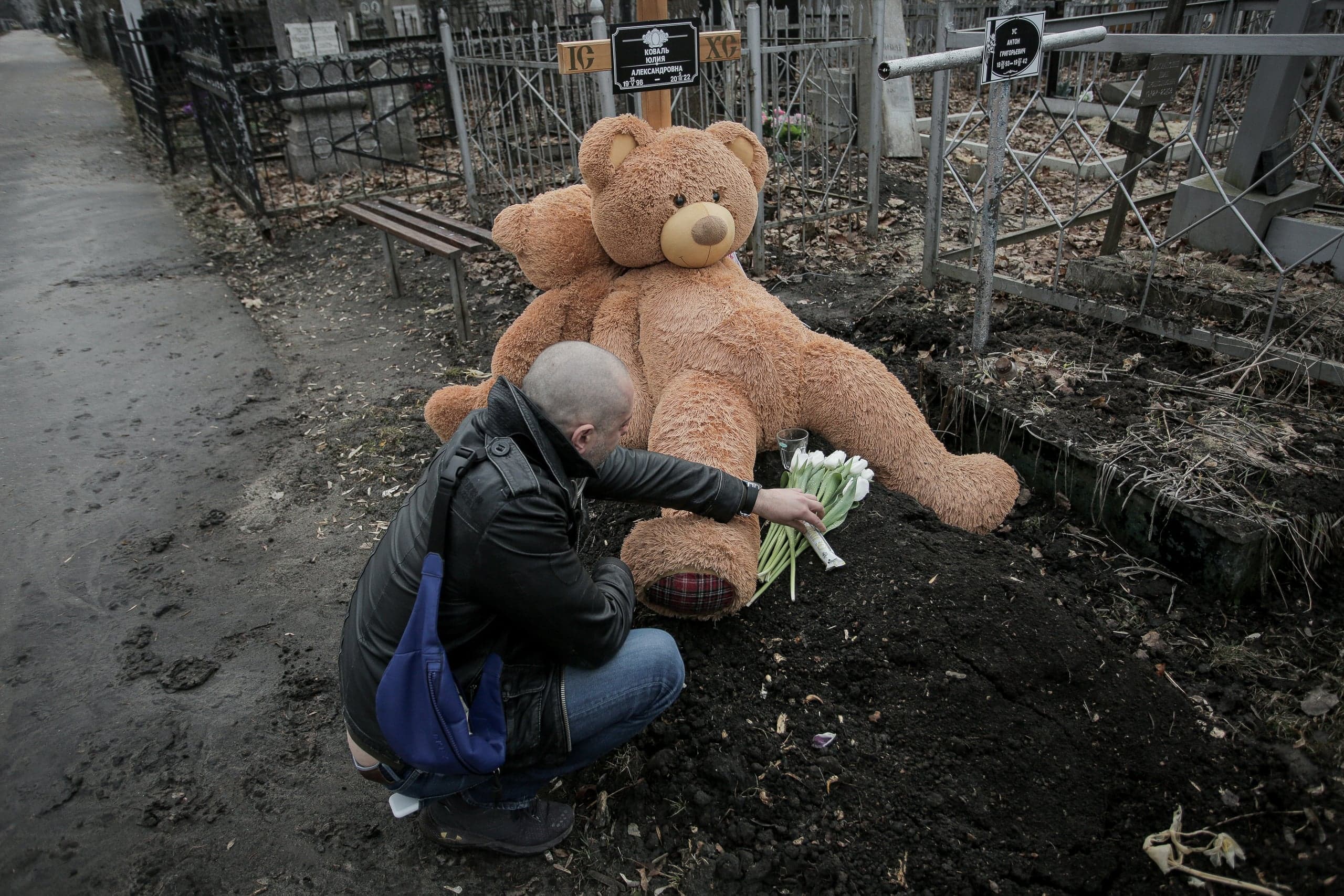 Мужчина кладет белые цветы на свежую могилу, украшенную большим плюшевым медведем. Надпись на кресте: "НАТАЛІЯ ОЛЕКСАНДРІВНА".