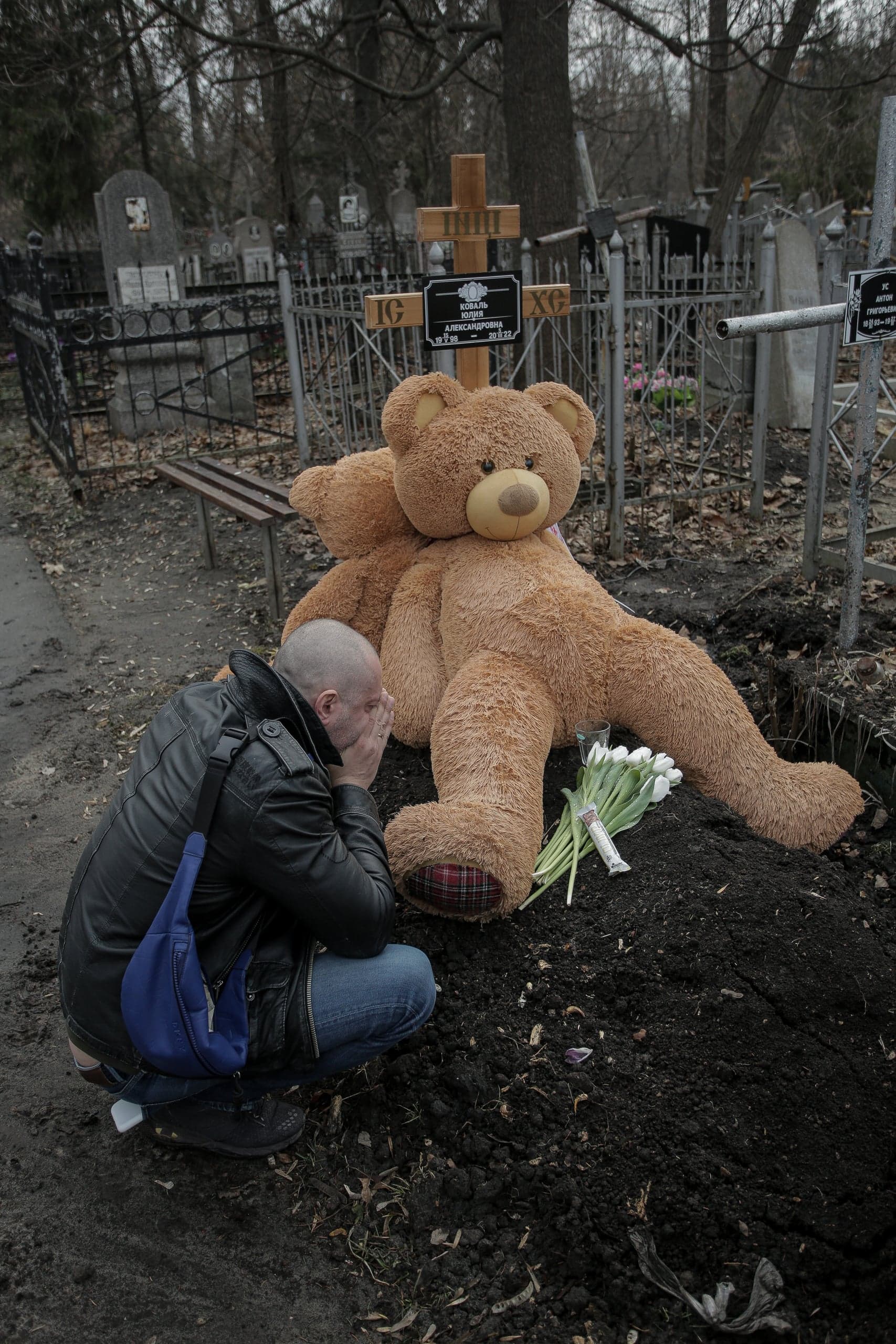 Мужчина скорбит на коленях у свежей могилы, на которой лежит гигантский плюшевый медведь и белые тюльпаны. На кресте надпись: «КОВАЛЬ ЮЛИЯ АЛЕКСАНДРОВНА».