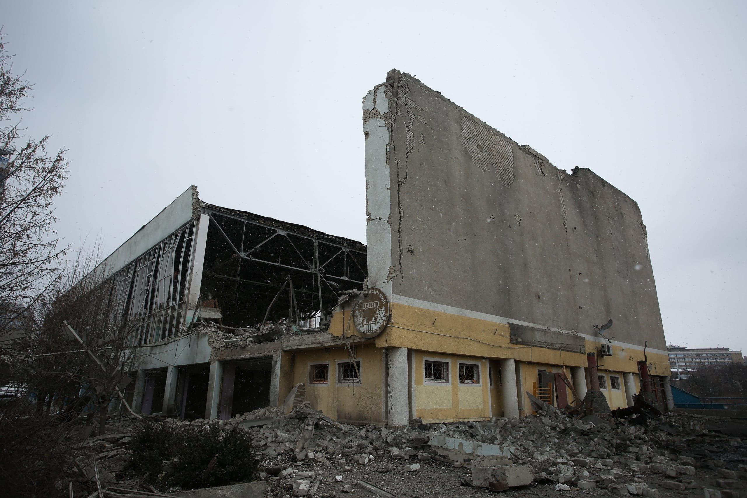 Фотография сильно разрушенного здания или спортивного комплекса, большая часть которого обрушилась, оставив стоять высокую голую фасадную стену. На переднем плане видны груды строительного мусора и обломков под пасмурным небом.
