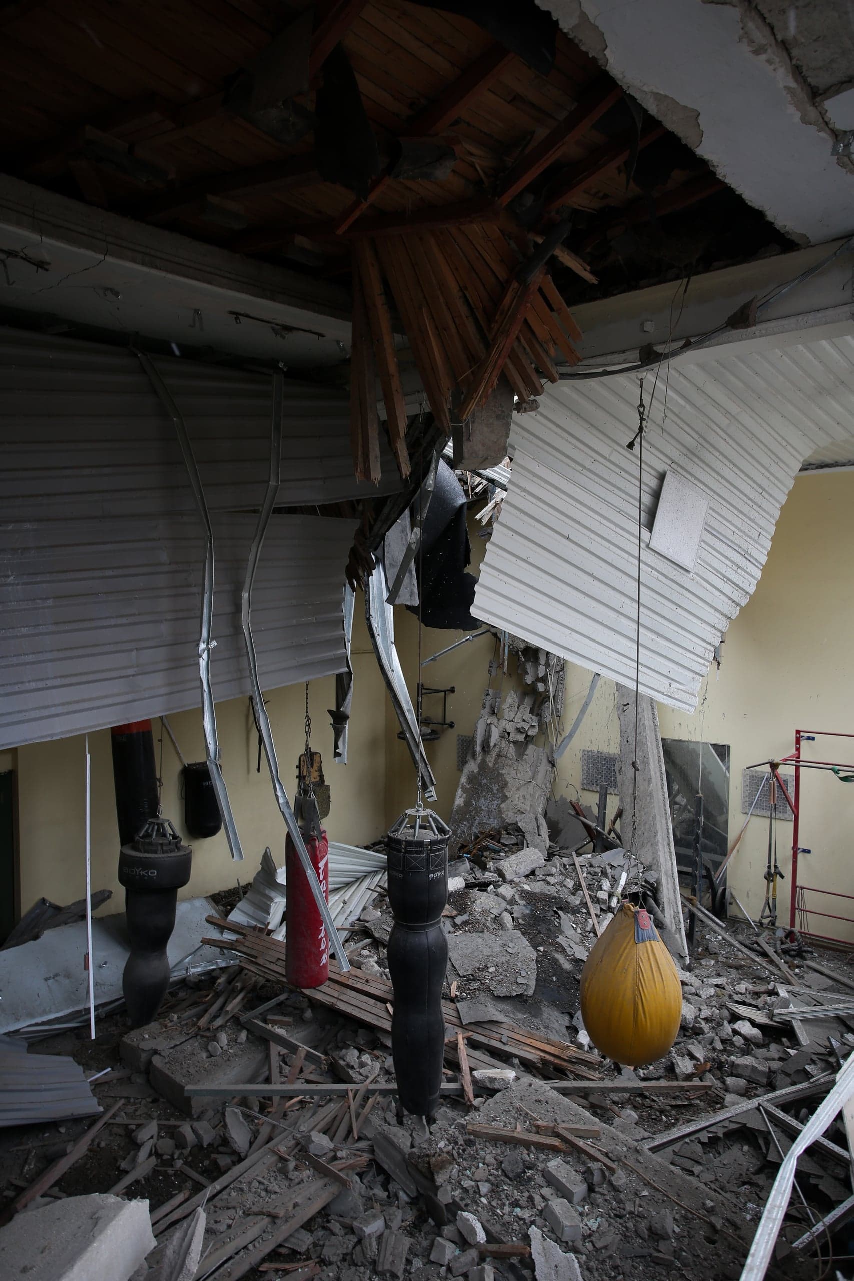 Интерьер сильно разрушенного боксерского зала, заваленного обломками бетона, дерева и металлического профиля. Частично обрушен потолок, а среди руин висят несколько боксерских груш.