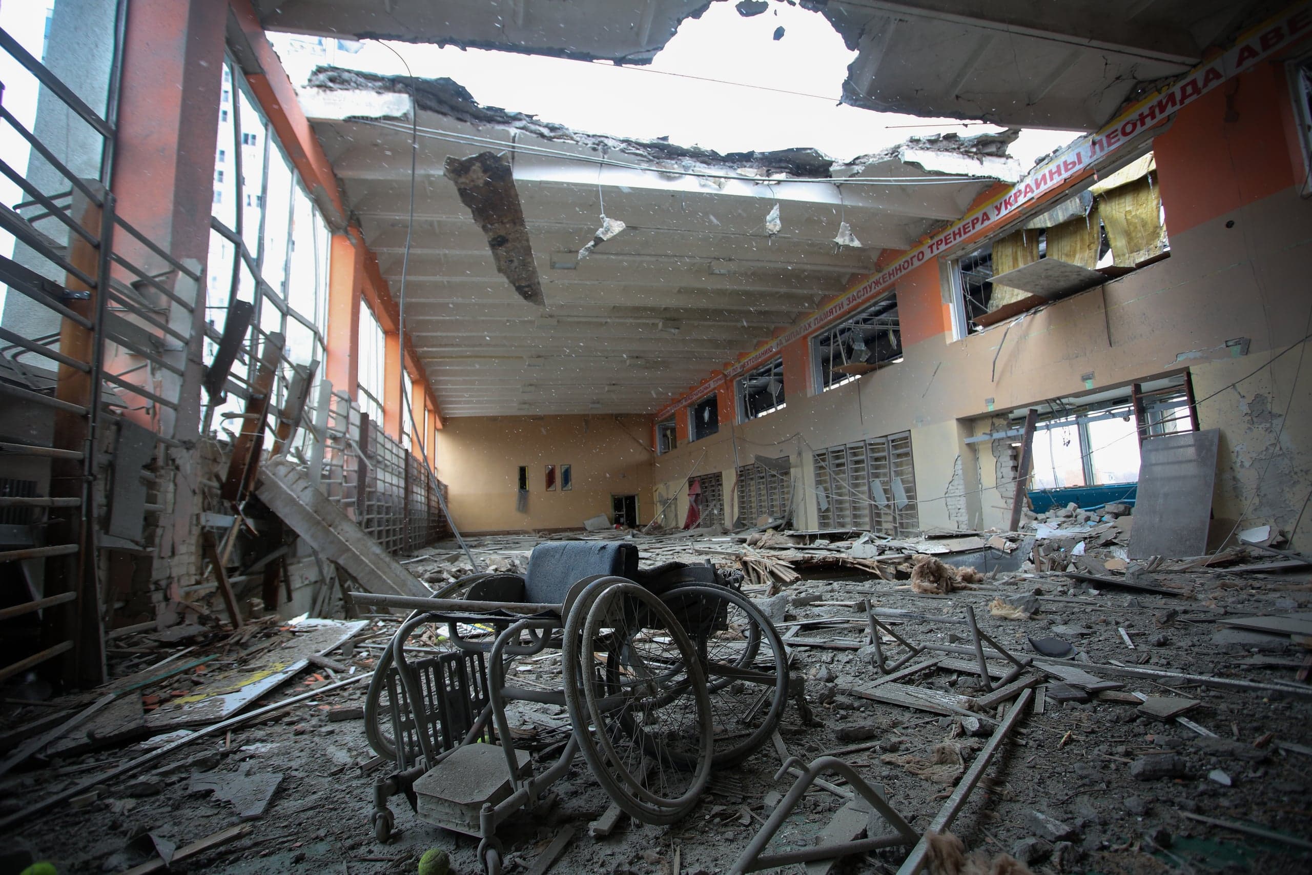 Крупный план разрушенного спортивного зала с пробоиной в крыше. На полу, покрытом обломками и пылью, лежит опрокинутое инвалидное кресло.