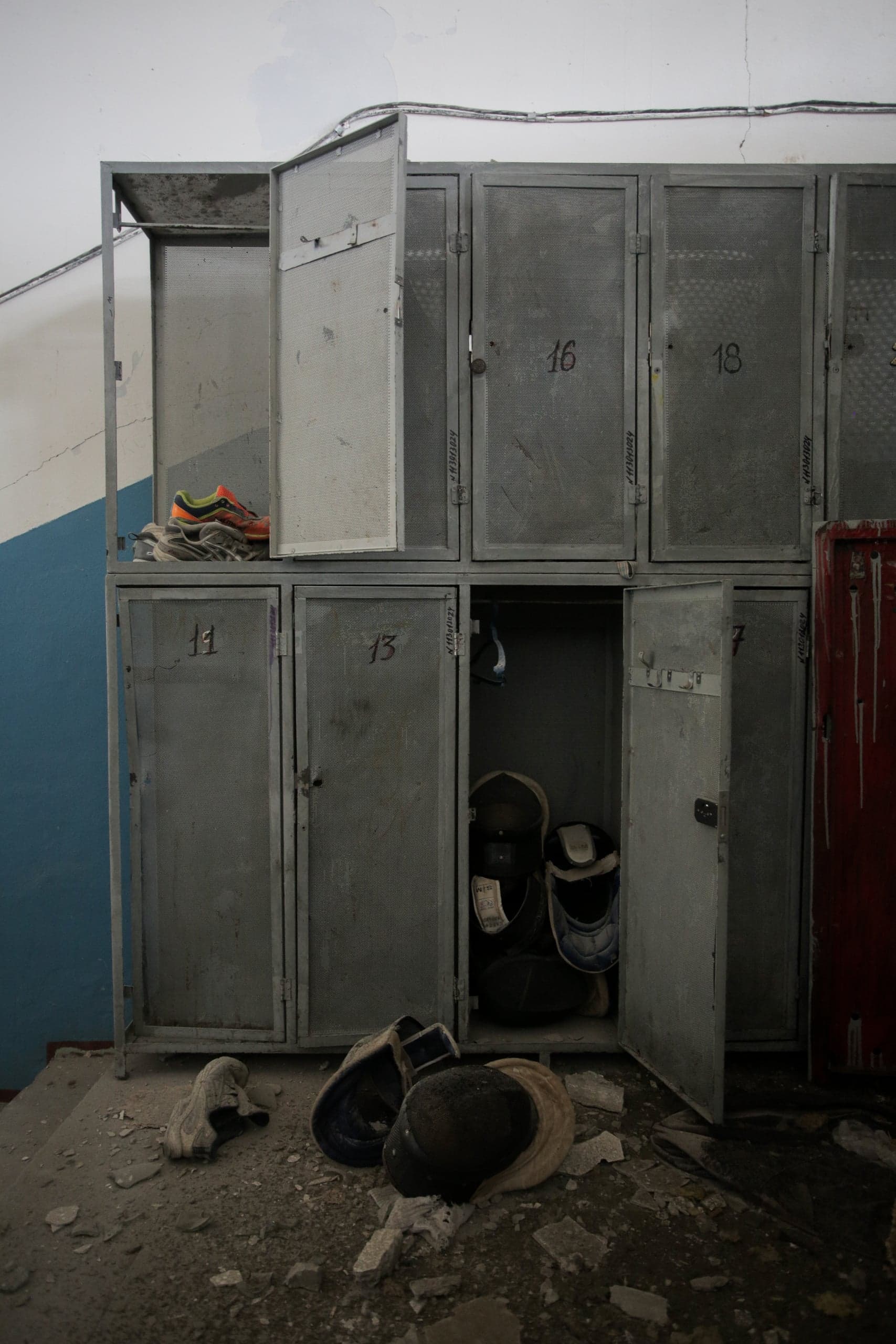 Ряд старых серых металлических шкафчиков с перфорированными дверцами в заброшенной, разрушенной комнате. Фехтовальные маски лежат на грязном полу среди обломков, а кроссовки — на верхней полке.