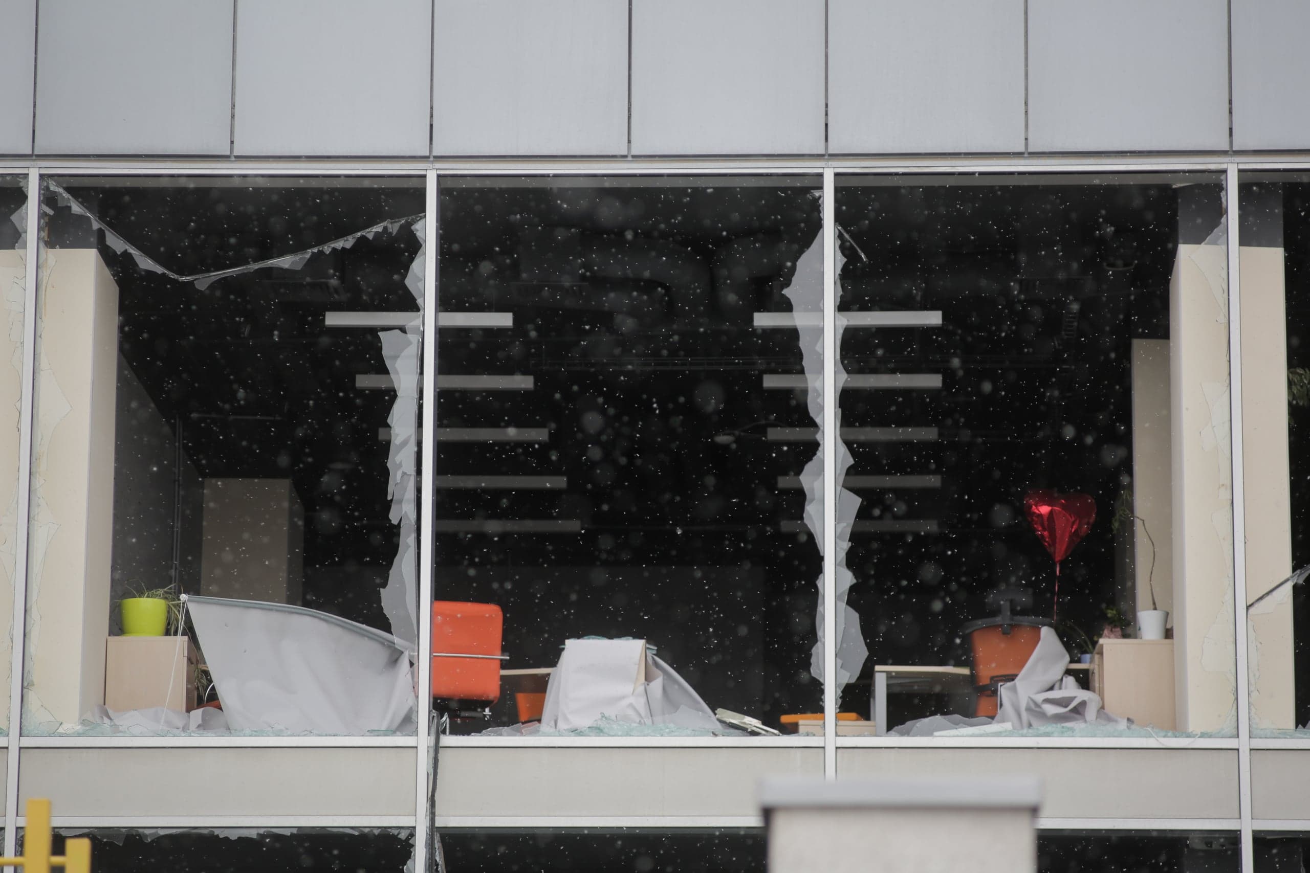 Разбитое панорамное окно современного здания, с крупными трещинами и свисающими осколками. Внутри видны офисная мебель, оранжевое кресло, накрытое тканью, и красный фольгированный шарик в форме сердца; сквозь стекло падают белые частицы, похожие на снег или пыль.