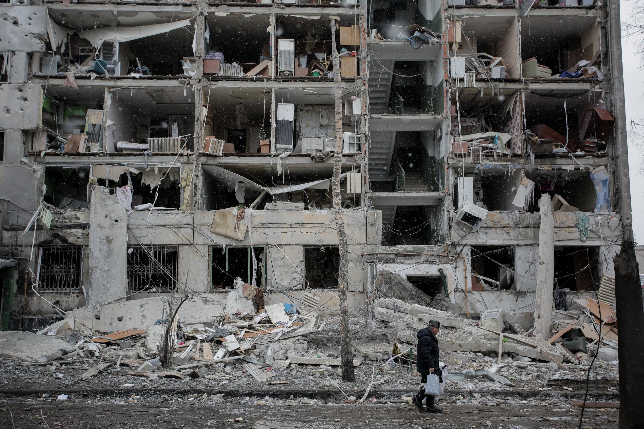 Крупный план сильно поврежденного многоэтажного жилого дома с полностью разрушенным фасадом. На фоне обнаженных внутренних комнат и лестничных пролетов сквозь падающий снег проходит человек в темной одежде.